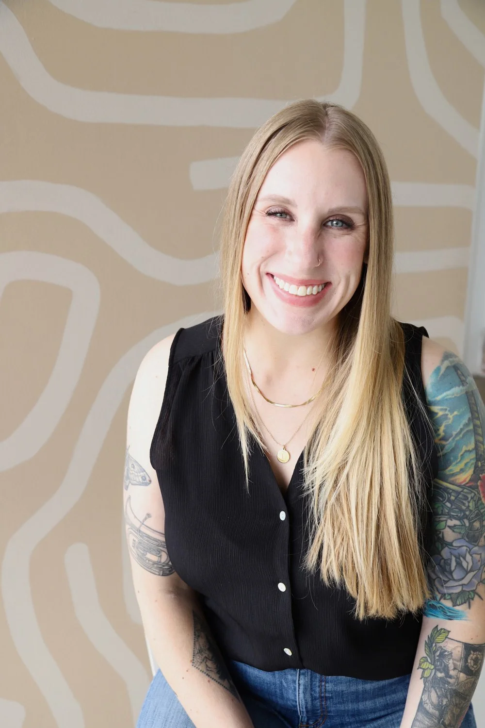 Lindsey is a freelance graphic designer specializing in brand + web design. She is a smiling woman with long blonde hair, tattoos on her arms, and wearing a black sleeveless top.