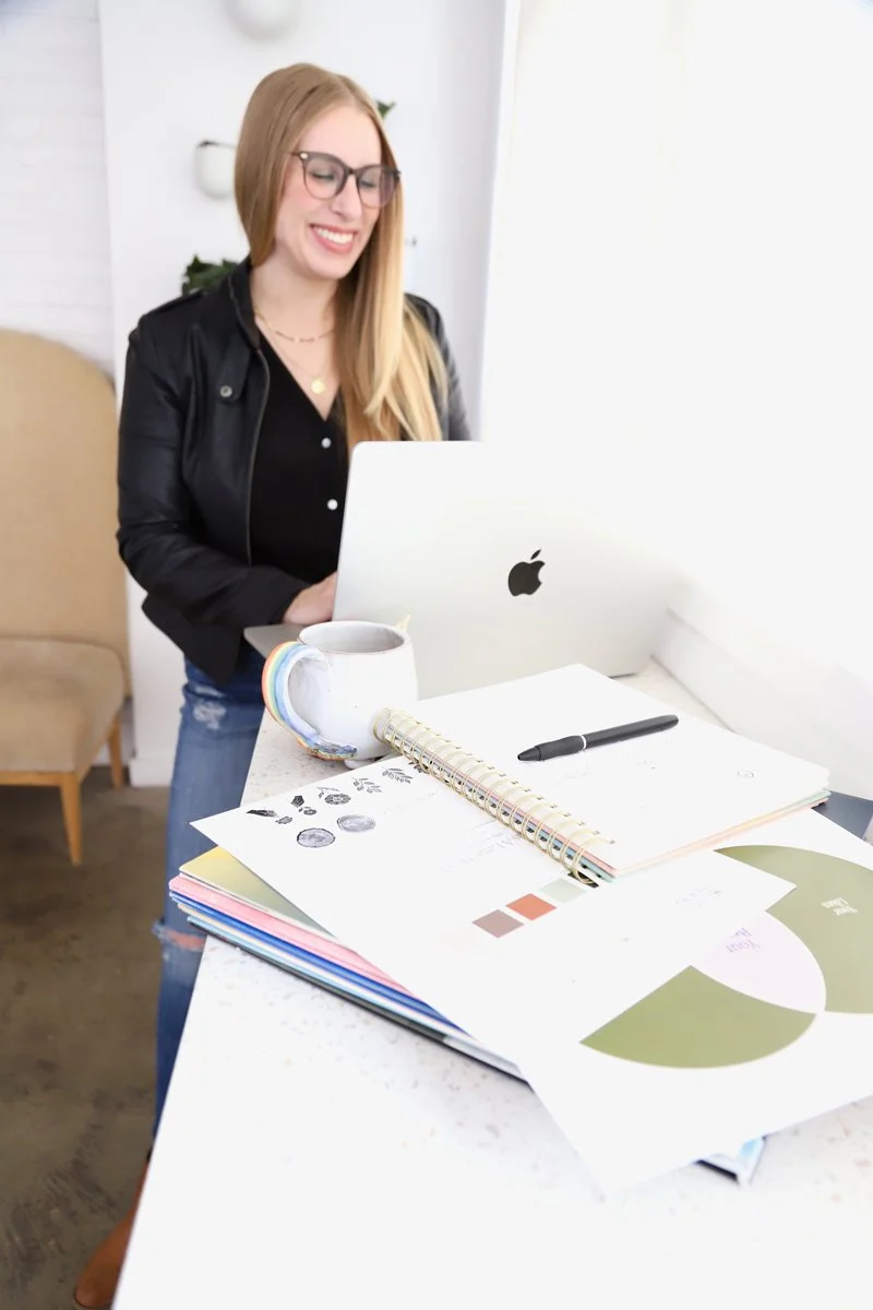 Lindsey is a freelance graphic designer specializing in brand + web design. She is a woman with long blond hair and glasses smiling while working on a laptop at a desk covered with papers, a black pen, a mug, and a spiral notebook.