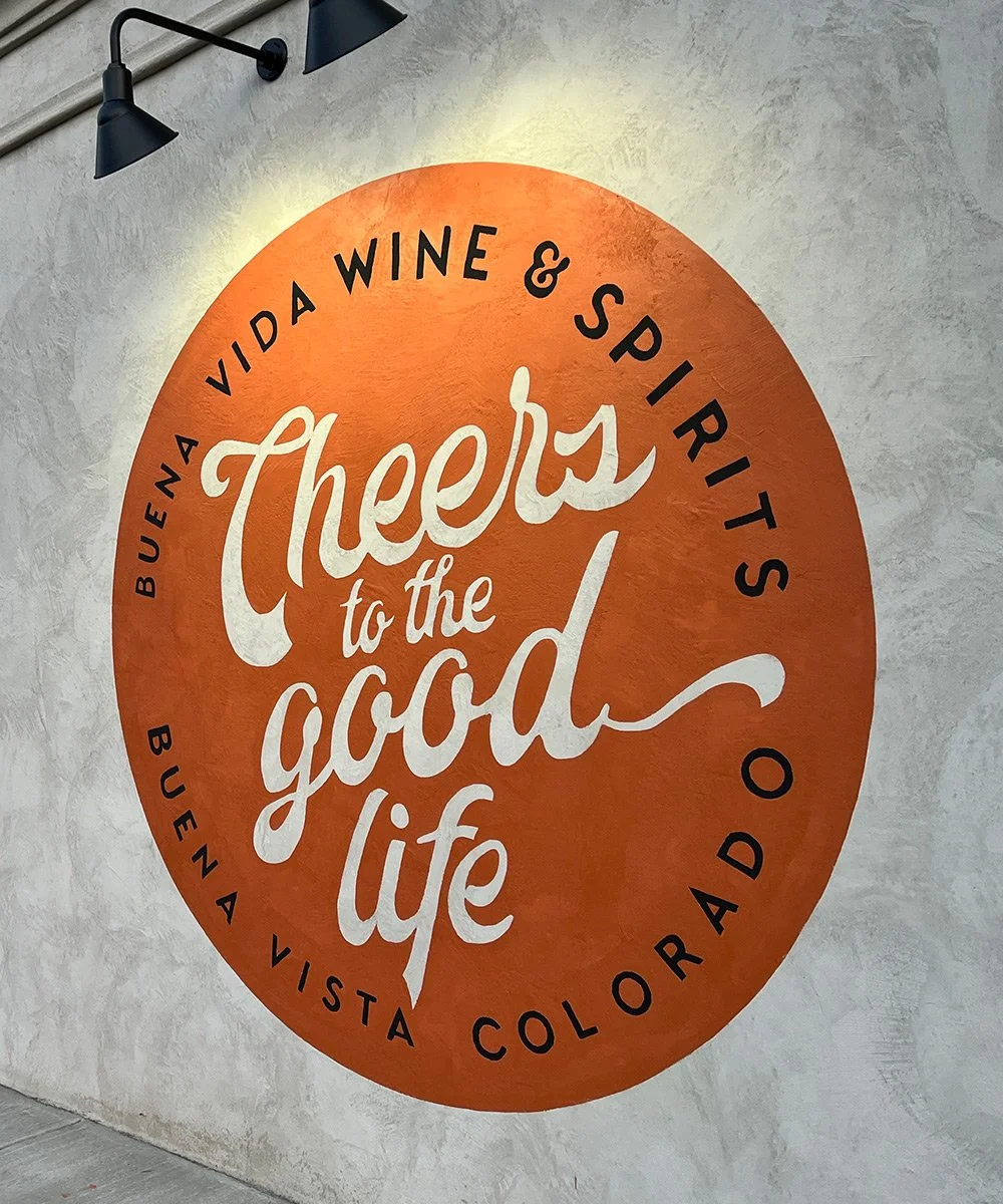 A circular logo design on a wall that reads 'Cheers to the good life' with the surrounding text 'Buena Vida Wine & Spirits' and 'Buena Vista Colorado'. There are black wall-mounted lamps above the sign.