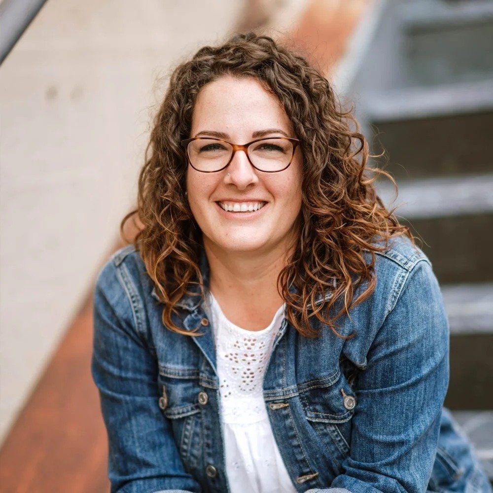 Hannah Dudley is a Six Leaf Design client and founder of Healthy Self. Healthy Life. She is a woman with glasses and curly brown hair smiling outdoors, sitting on steps, wearing a denim jacket and white top.