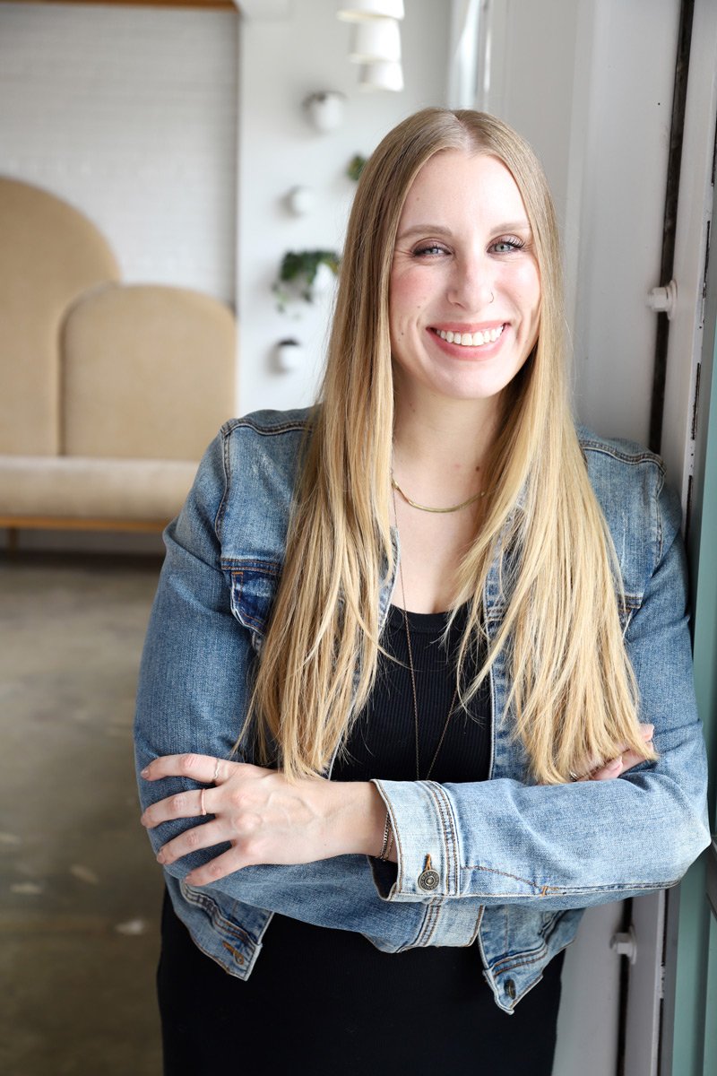Lindsey is a freelance graphic designer specializing in brand + web design. She is a smiling woman with long blonde hair, wearing a denim jacket and black top, standing near a window in a brightly lit room.