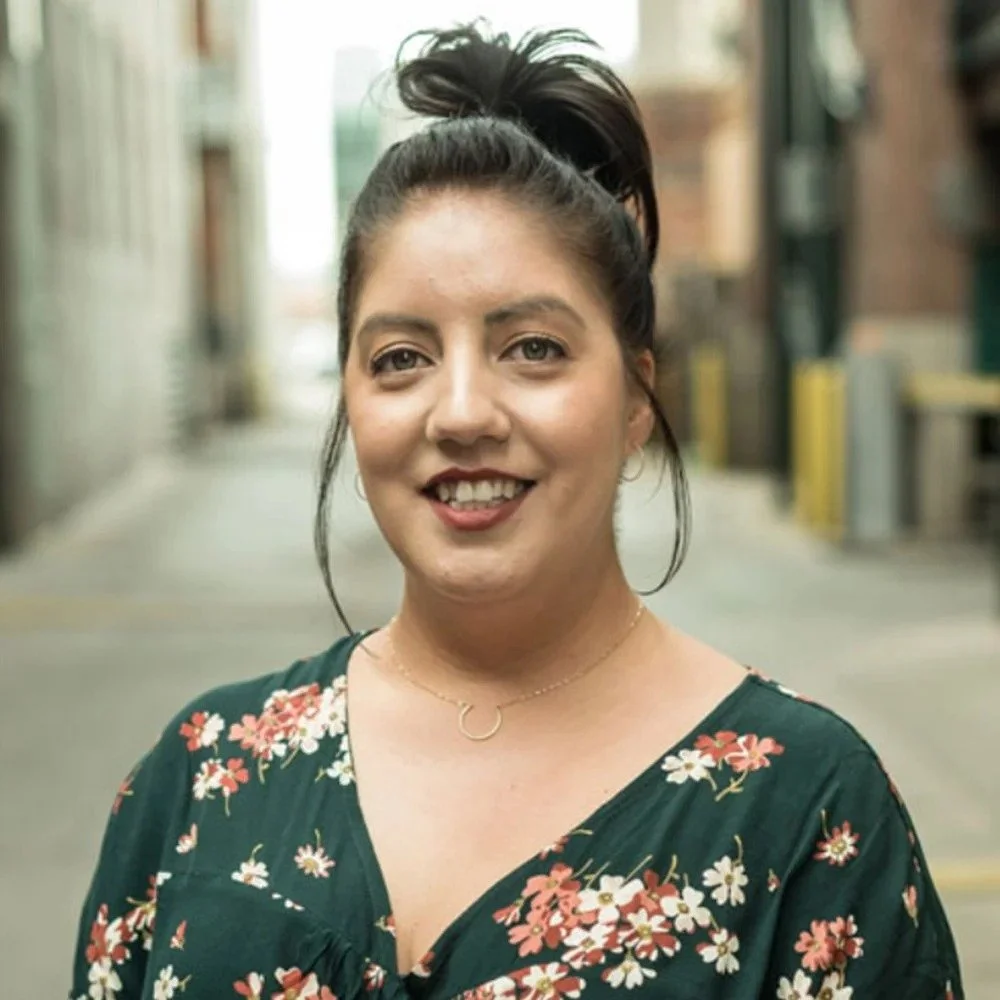 Lucy Taylor is a Six Leaf Design client and marketing manager of Brightstores. She is a woman with long dark hair tied up in a high bun, smiling at the camera, standing outdoors on a city street, wearing a dark green floral dress and a gold necklace.