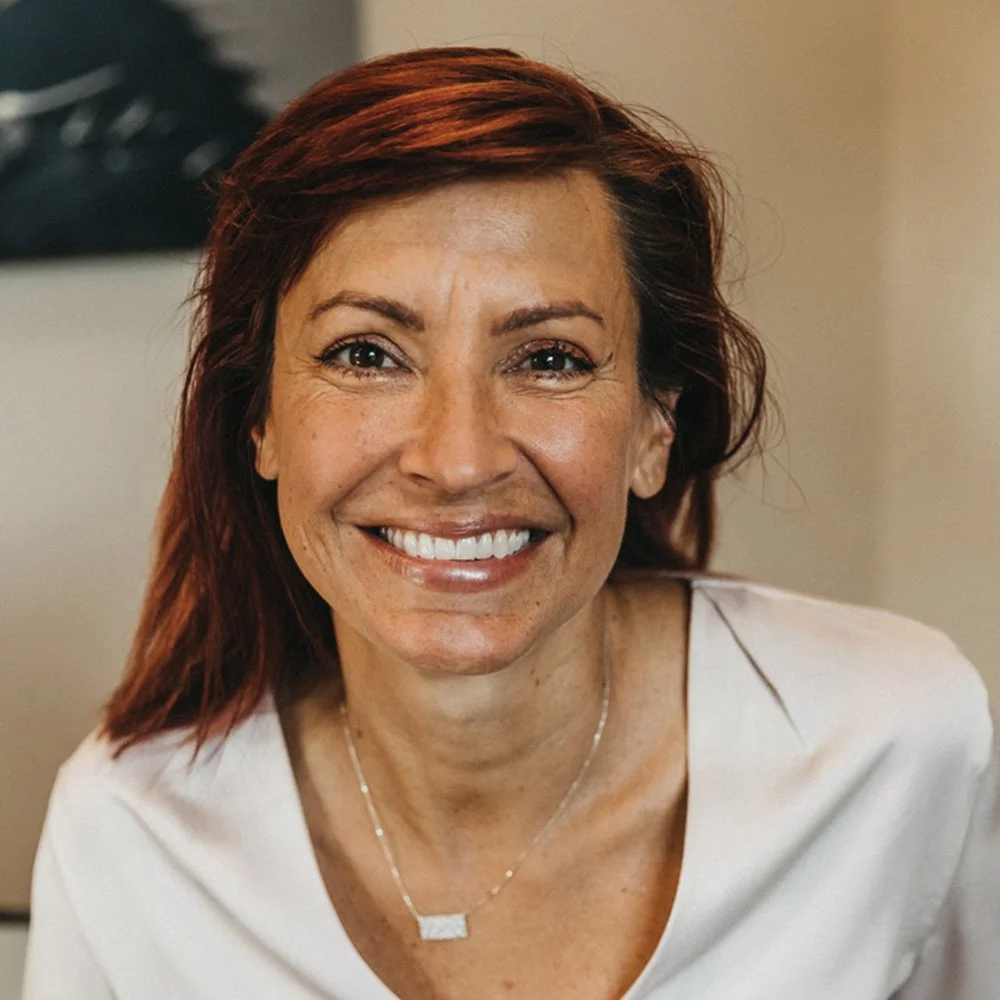 Lara Postma is a Six Leaf Design client and founder of The Great Relate. She is a smiling woman with shoulder-length reddish-brown hair, wearing a white top and a delicate necklace, sitting indoors with a blurred background.