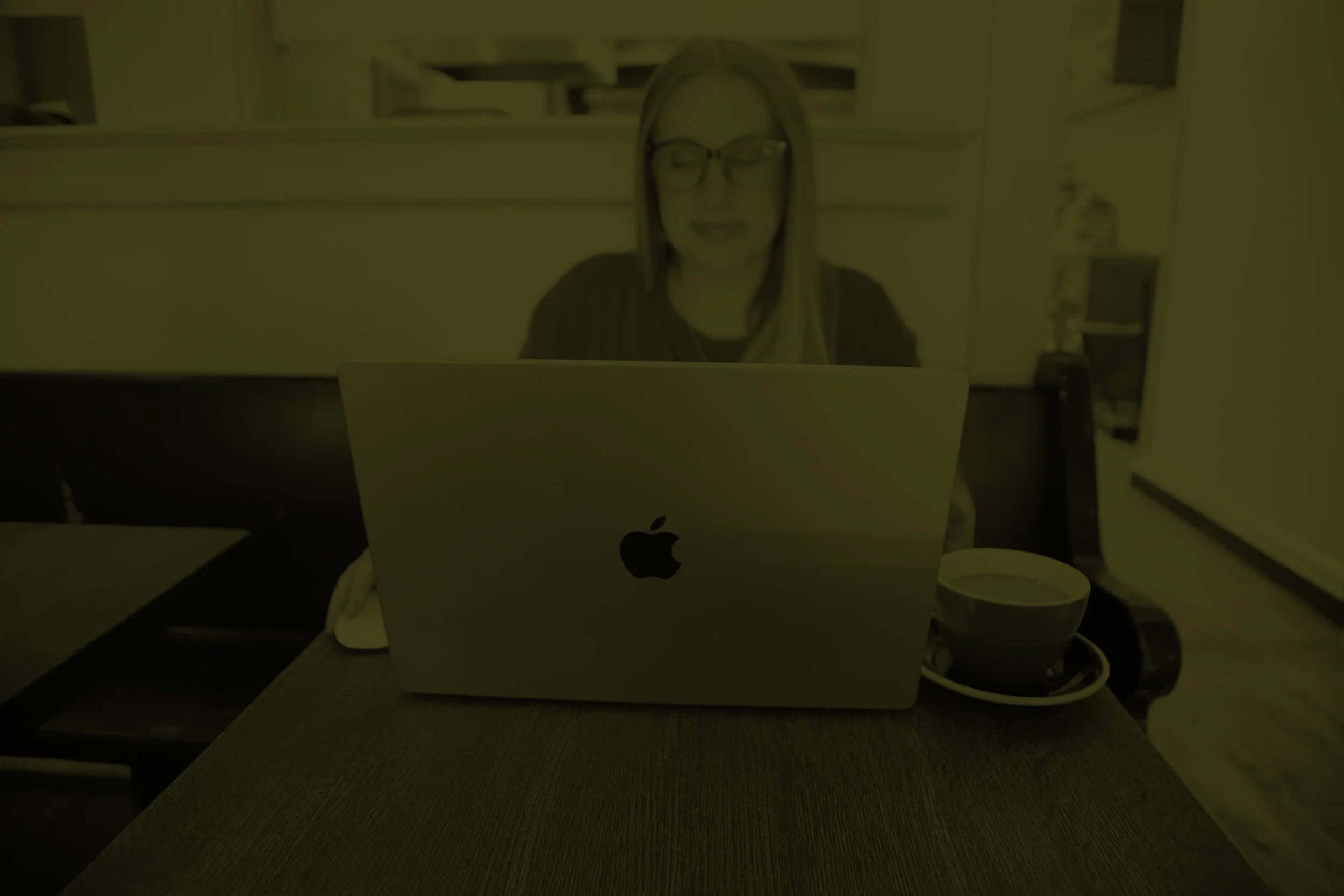 Lindsey is a freelance graphic designer specializing in brand + web design. She is a woman with glasses sitting at a table with a silver MacBook and a cup of coffee.