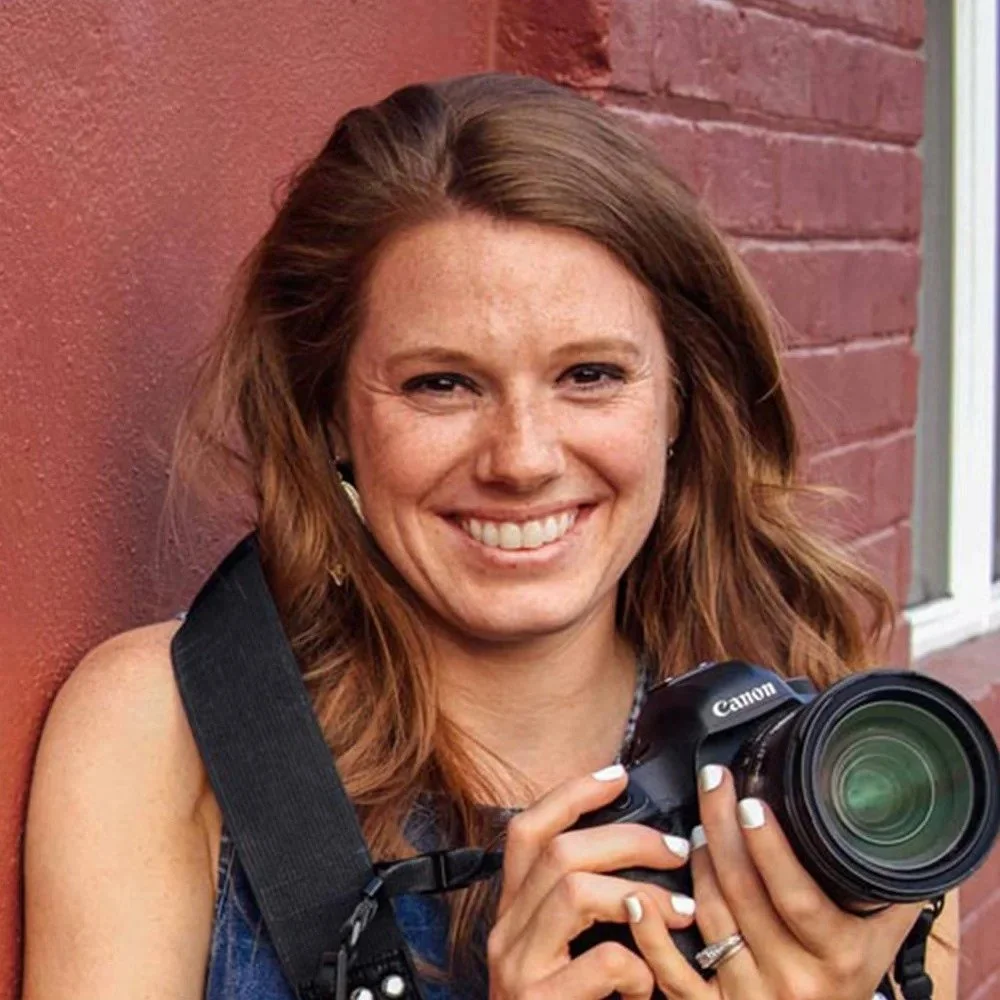 Rebecca Todd is a Six Leaf Design client and owner of TruBlue Images. She is a woman holding a Canon camera, smiling, leaning against a brick wall.
