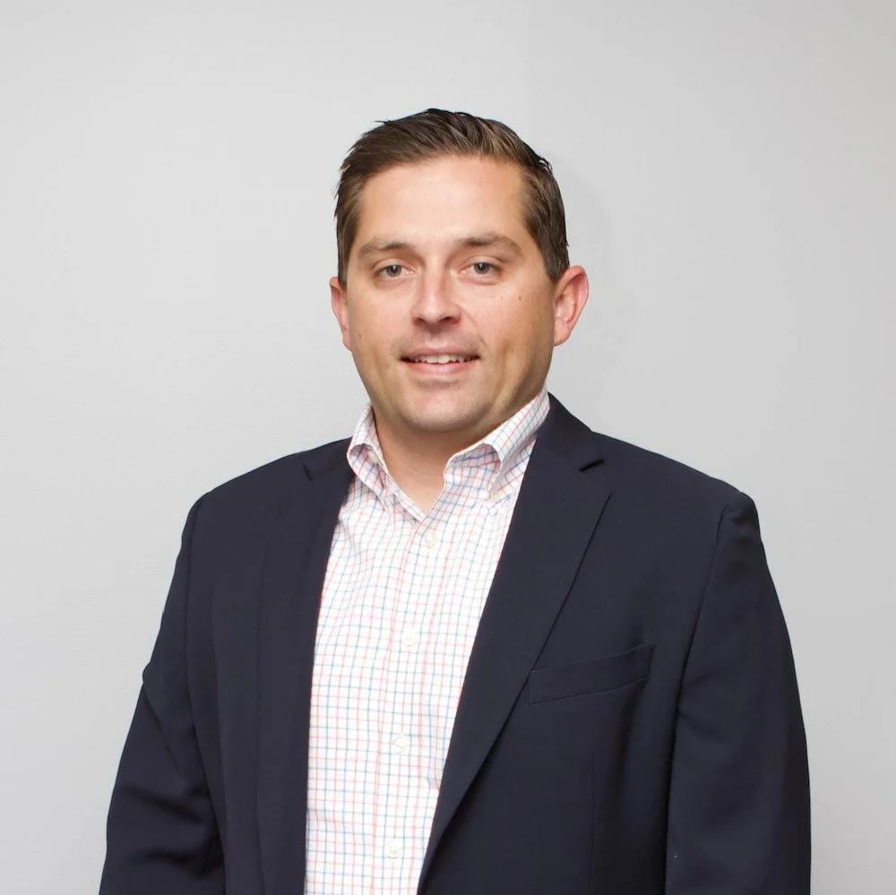 John Sutton is a Six Leaf Design client and Pricipal of Newpoint Services. He is a professional man in a dark suit and checkered shirt standing against a plain white wall.