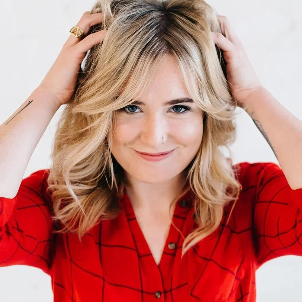 Emily Weinkauf is a Six Leaf Design client and owner of Cedar Hair Studio. She is a young woman with blonde hair, blue eyes, wearing a red plaid shirt, smiling, holding her hair with both hands, against a plain white background.