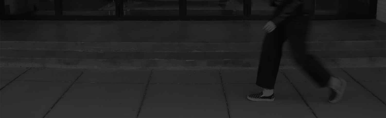 A brand, print, + web designer walking past a building entrance with stairs. The person is wearing dark pants, checkered slip-on shoes, and a dark jacket. The image is in black and white.