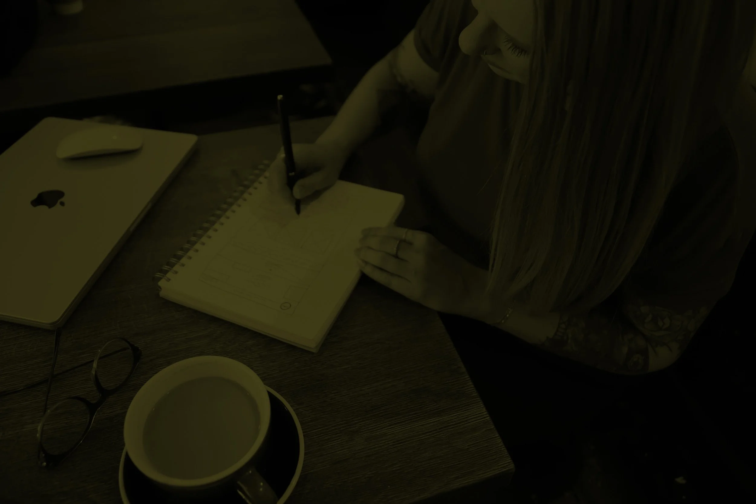 Brand + website designer writing in a notebook with an Apple laptop, eyeglasses, and a coffee cup on a wooden table.
