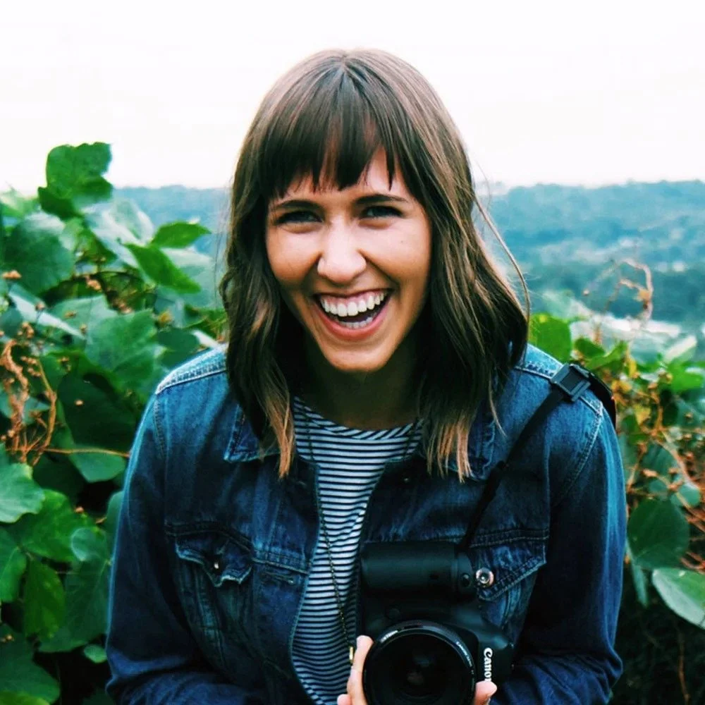 Alexandra Davie is a Six Leaf Design client and owner of Alexandra Davie Photography. She is a young woman with brown hair and bangs, smiling and wearing a denim jacket and striped shirt, holding a camera in a lush, green outdoor setting.