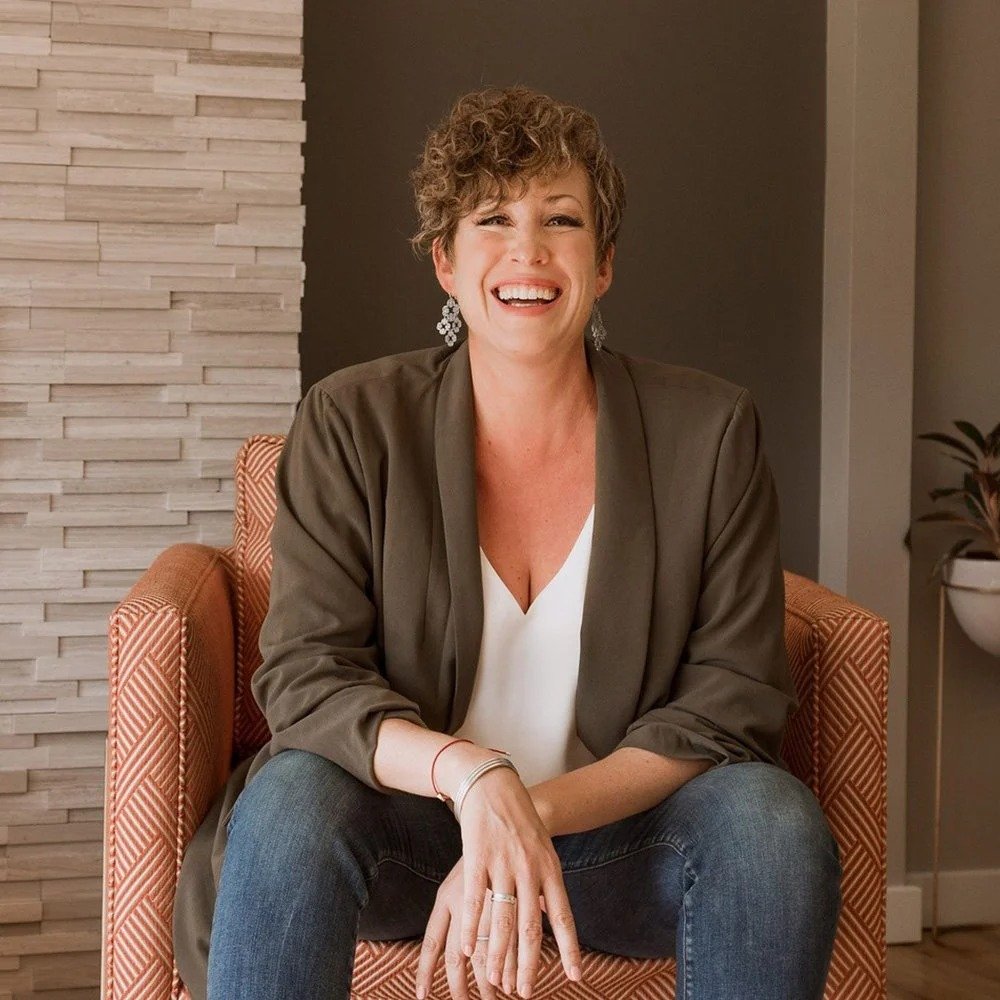 Lisa Kuzman is a Six Leaf Design client and a trauma-informed coach. She is a woman with short curly hair, smiling, sitting on an orange patterned chair in a modern room. She is wearing a white top, olive green blazer, and blue jeans.