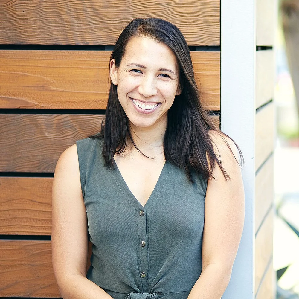 Courtney Shen Deshelter is a Six Leaf Design client and owner of SoCal Couples Therapy. She is a woman with dark brown hair and light skin smiling, wearing a sleeveless green top, standing outdoors against a wooden wall.