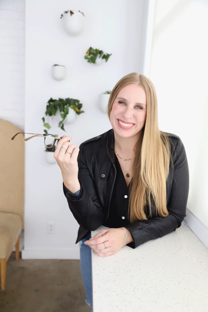 Lindsey is a freelance graphic designer specializing in brand + web design. She is a woman with long blonde hair, smiling, holding eyeglasses, sitting by a white countertop in a bright, modern room with white walls and decorative plants.