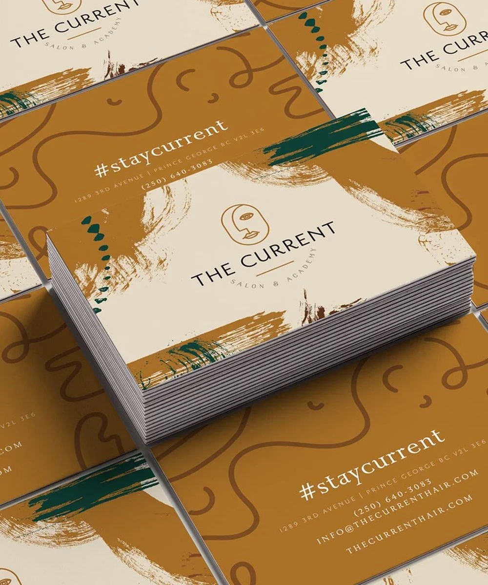 Business card design for The Current Salon & Academy with abstract gold and cream design, logo of a stylized face, and contact information including the hashtag #staycurrent and address in Prince George, BC.
