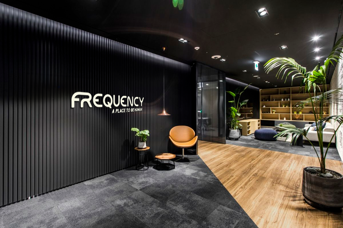 Modern office lounge area with black textured wall, white sign reading "FREQUENCY: A PLACE TO BE HUMAN," and stylish furniture including a tan chair, small table, and large potted plants.