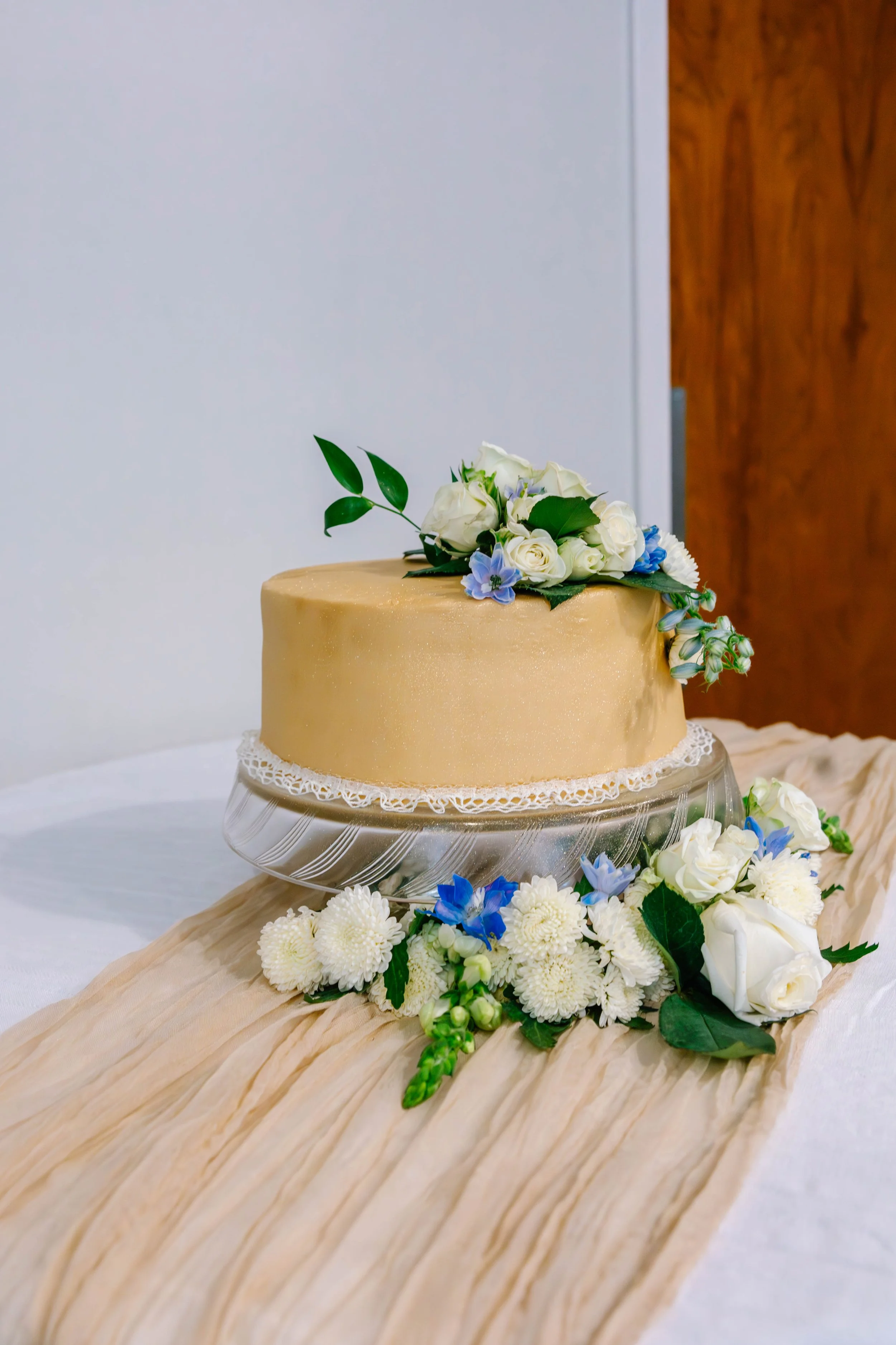 A decorated wedding cake with white roses, blue flowers, and green leaves, placed on a glass cake stand, surrounded by matching flowers on a beige cloth.