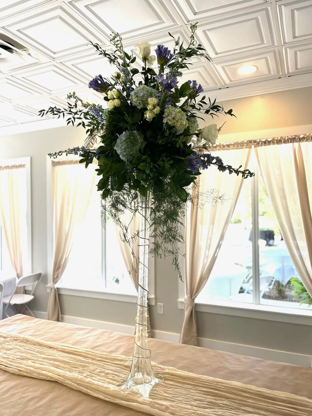 A tall floral centerpiece with white, purple, and green flowers on a table, set in a clear glass vase, in a room with large windows and beige curtains.