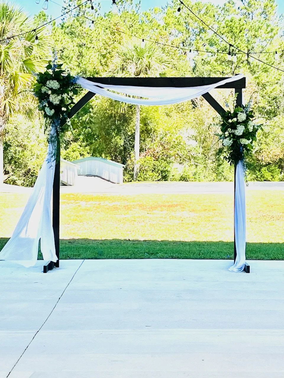 Outdoor wedding arch decorated with white and dark green flowers, white and black drapes, under string lights, on a patio with trees in the background.