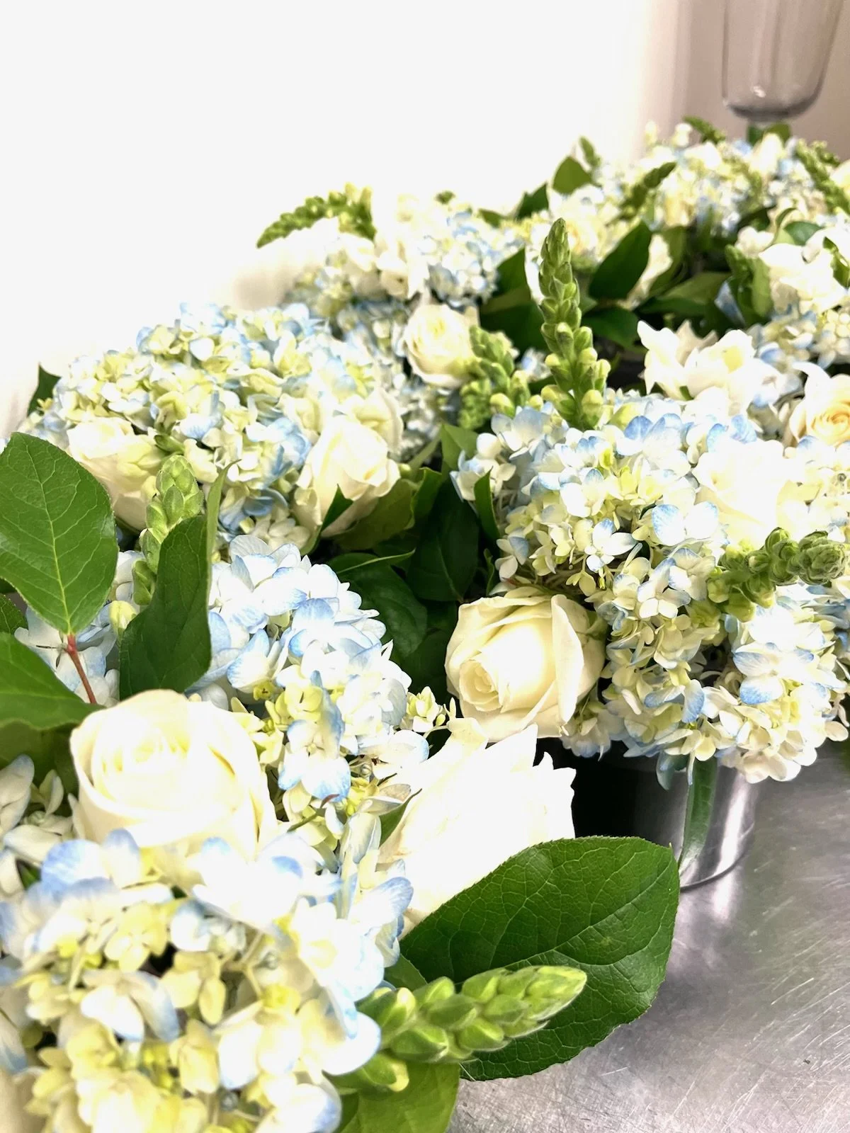 A close-up of a floral arrangement featuring white roses, light blue hydrangeas, green leaves, and other flowers in metal containers.