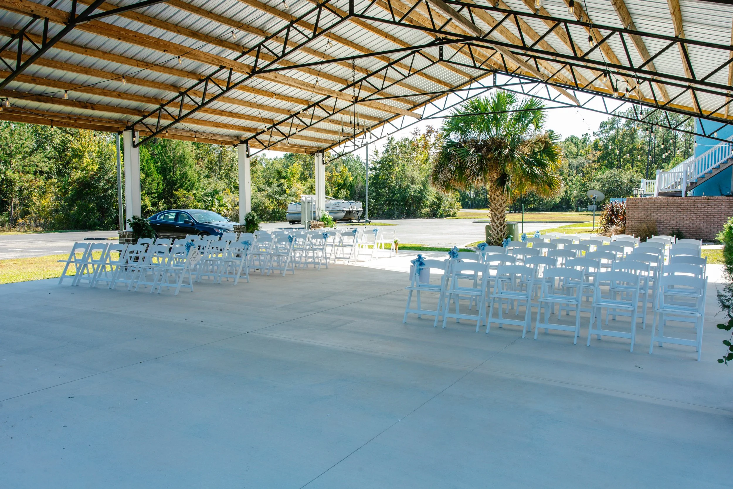 Outdoor wedding ceremony setup with white chairs decorated with blue ribbons under a metal roof pavilion, with palm trees and greenery in the background.