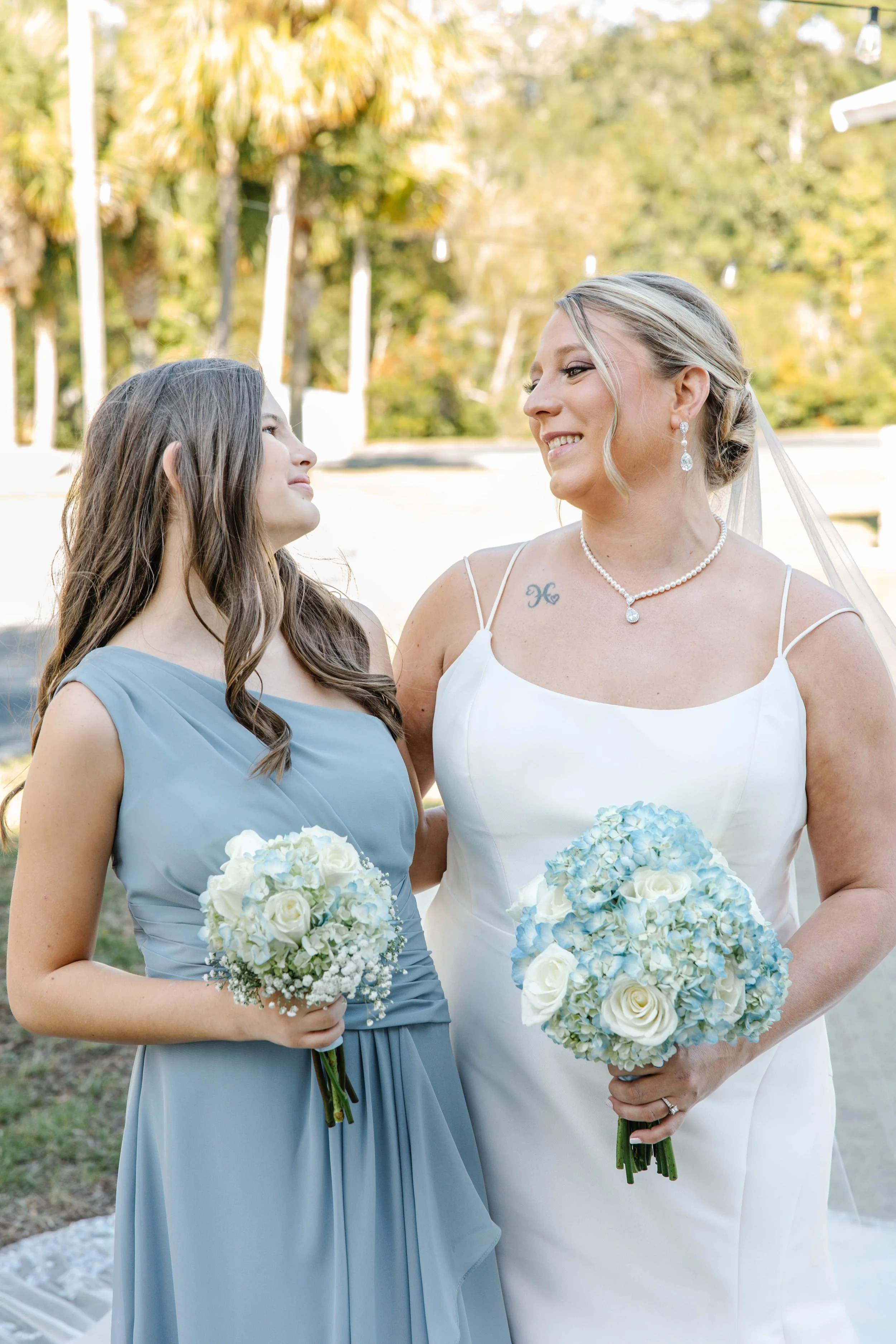 A bride and a bridesmaid during a wedding ceremony outdoors, holding bouquets of white roses, with the bride wearing a white dress and the bridesmaid in a light blue dress, smiling at each other.