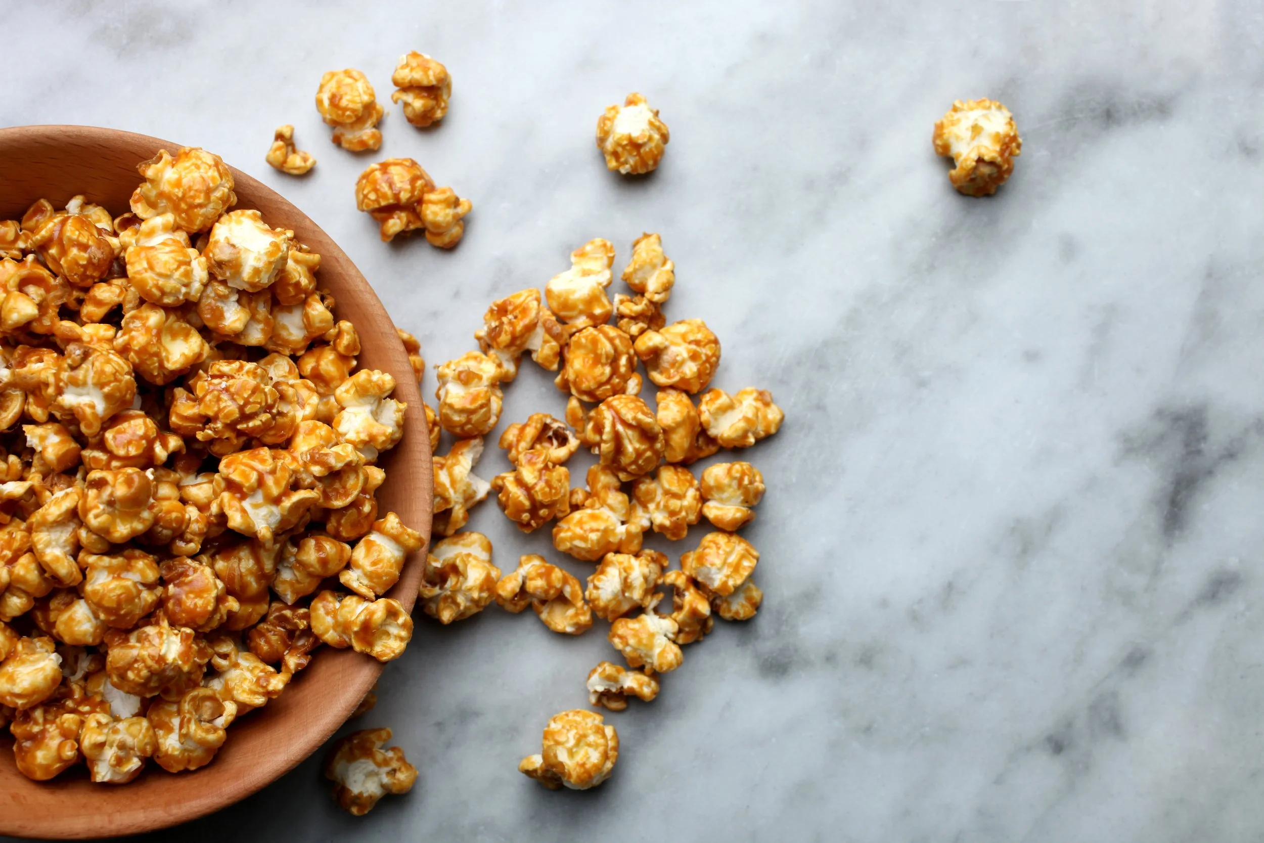 A wooden bowl filled with caramel popcorn on a marble surface, with some popcorn spilled outside the bowl.