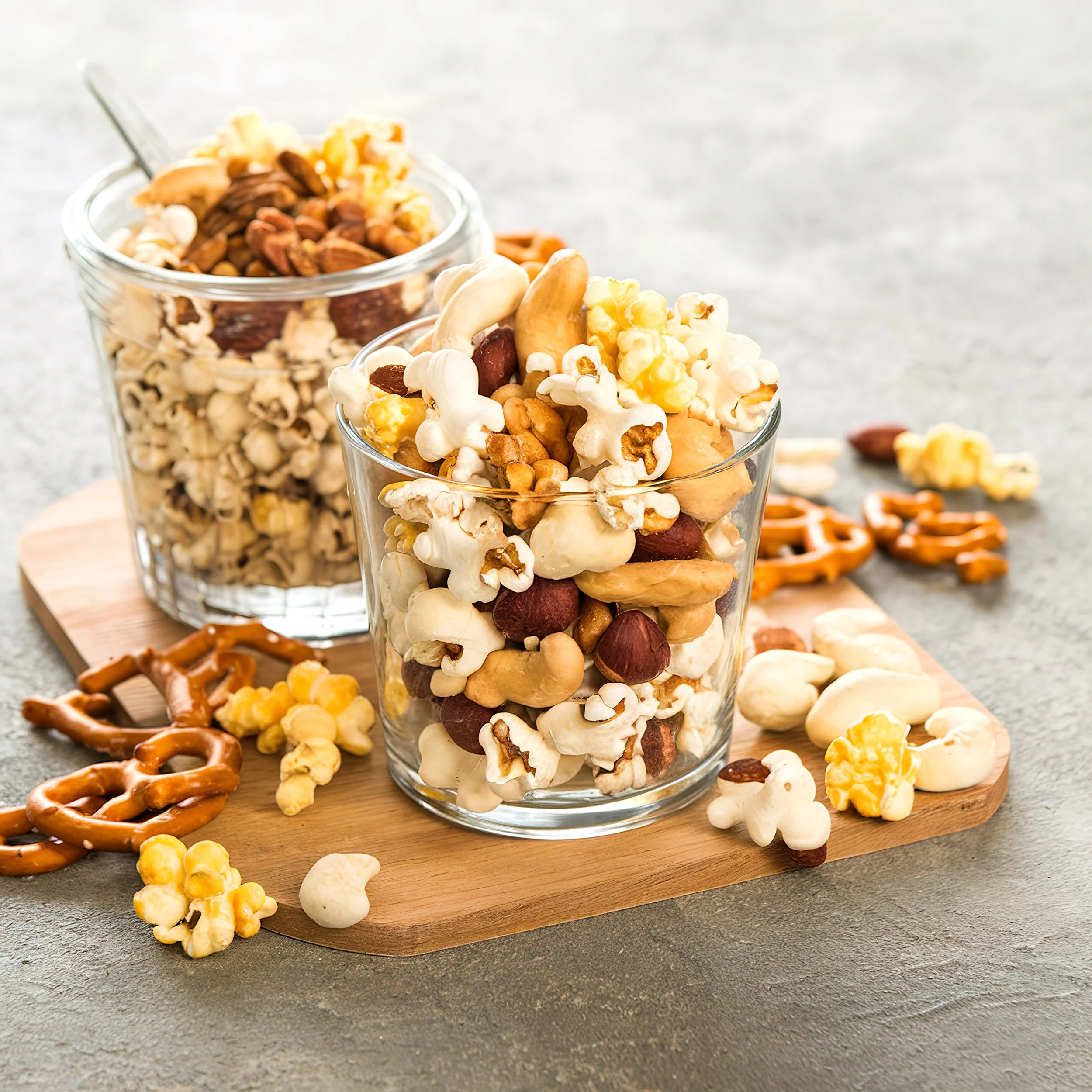 Two glasses of assorted mixed nuts, popcorn, and pretzels on a wooden board with some pieces spilled around.