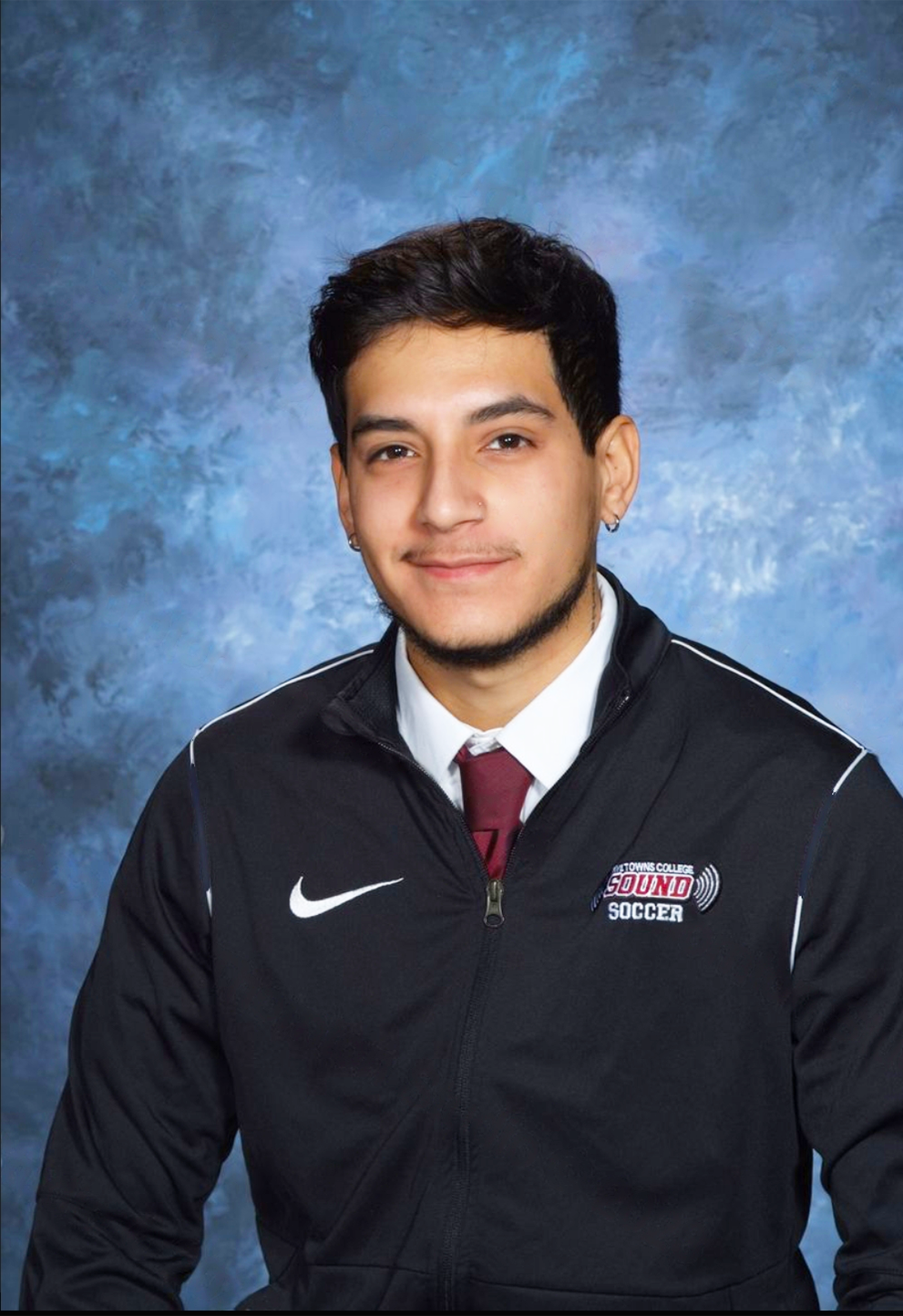 A young man with dark hair, facial hair, wearing earrings, a white shirt, maroon tie, and a black Nike jacket with a 'Guilford College Sound Soccer' logo, posing against a blue textured background.