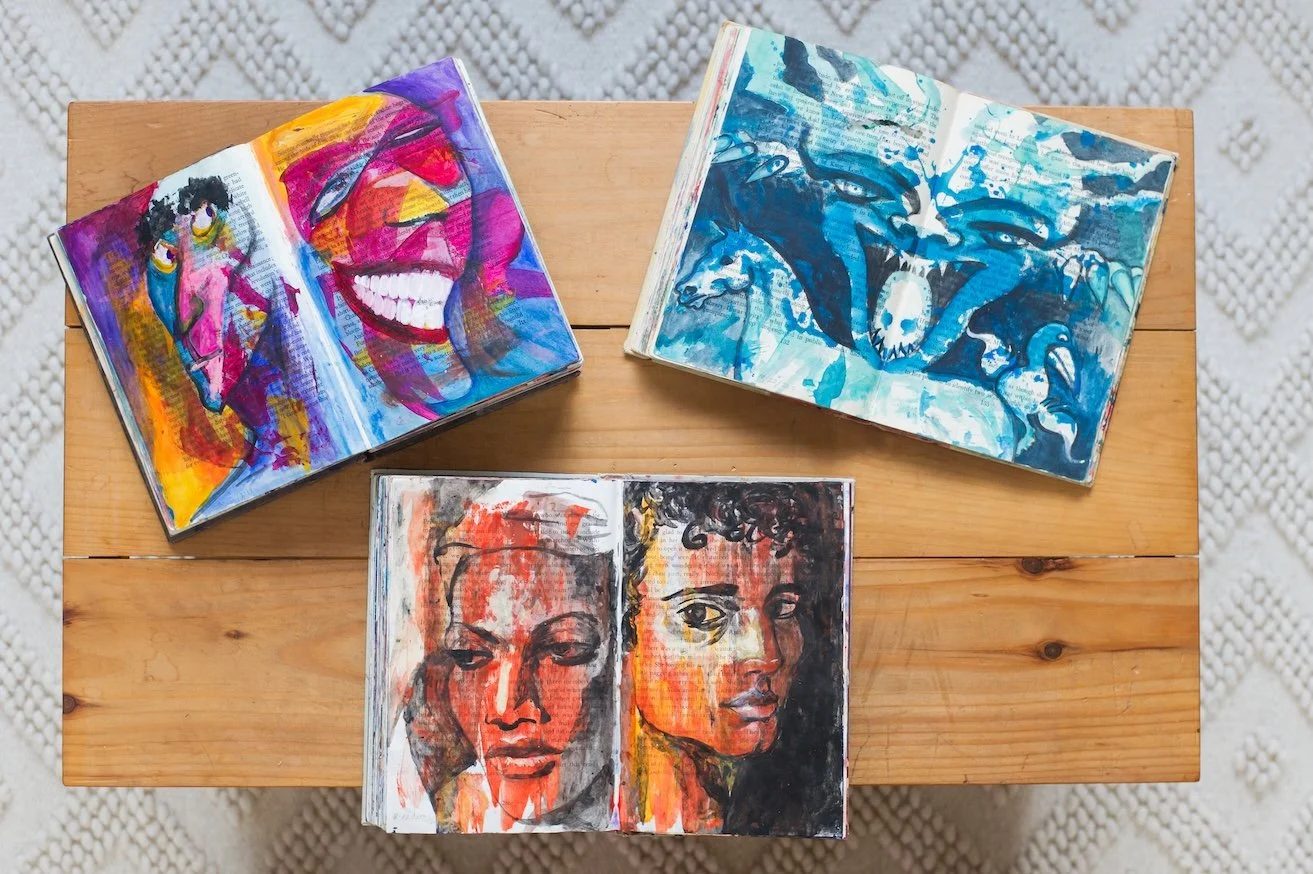 Three open sketchbooks with colorful portraits and illustrations on a wooden table.