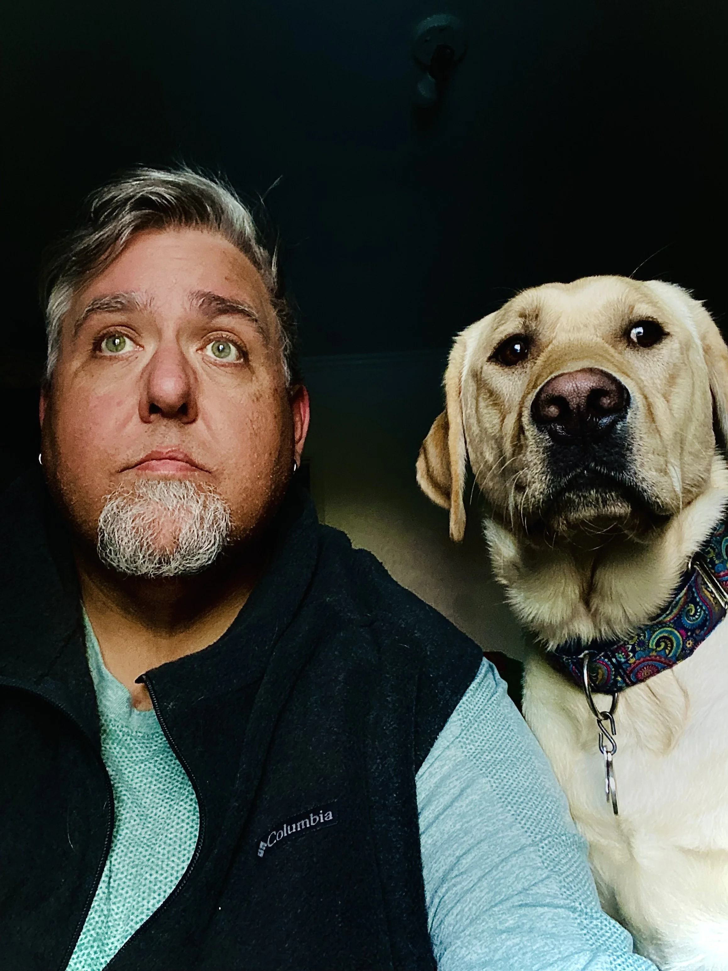 A man with gray hair and a beard taking a selfie with a yellow Labrador Retriever dog. The man has green eyes and is wearing a black Columbia vest over a teal shirt. The dog is wearing a colorful bandana collar and has a calm expression.