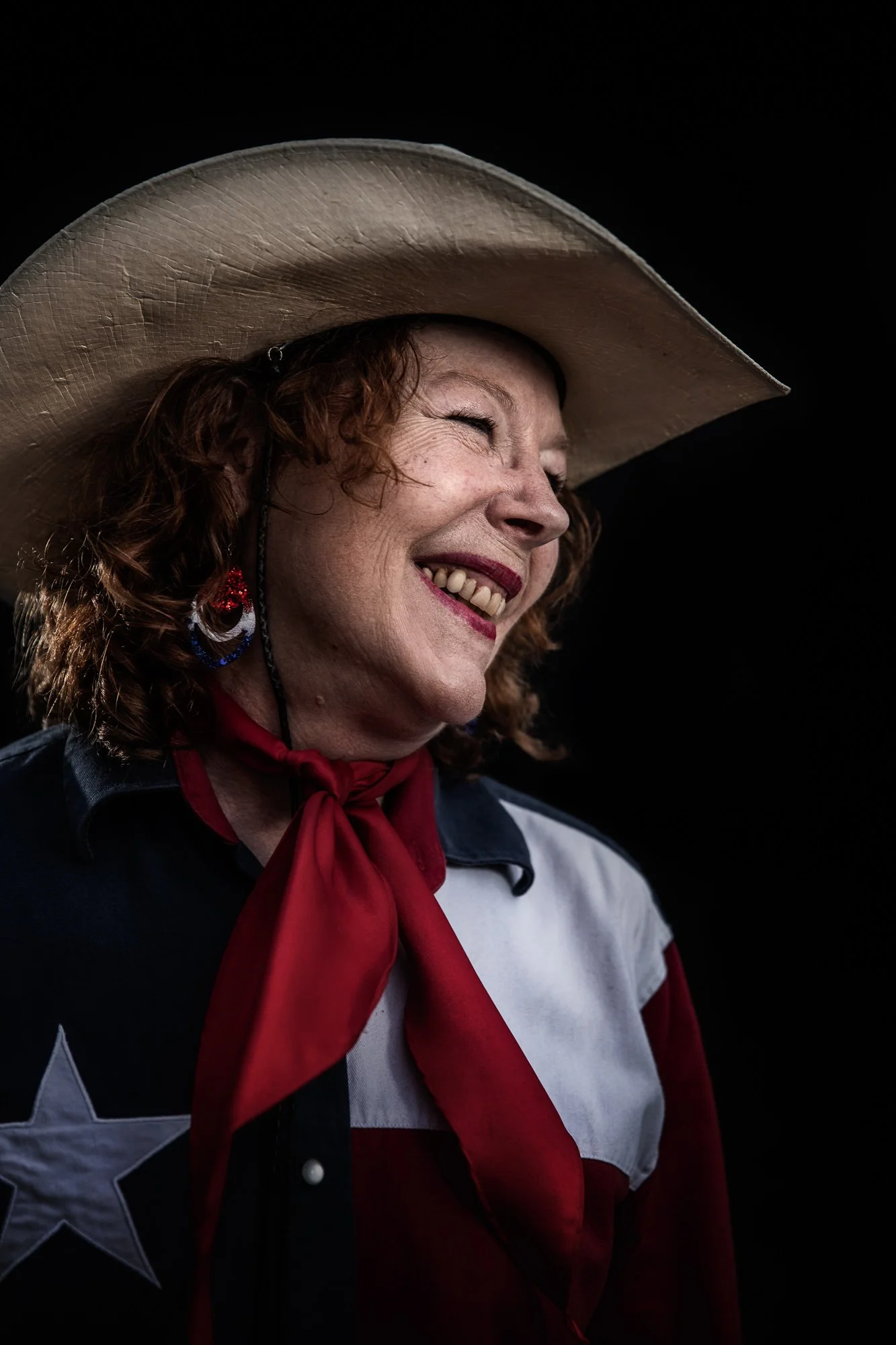 A woman with curly red hair wearing a cowboy hat, red bandana, and a shirt with stars, smiling and laughing with her eyes closed.