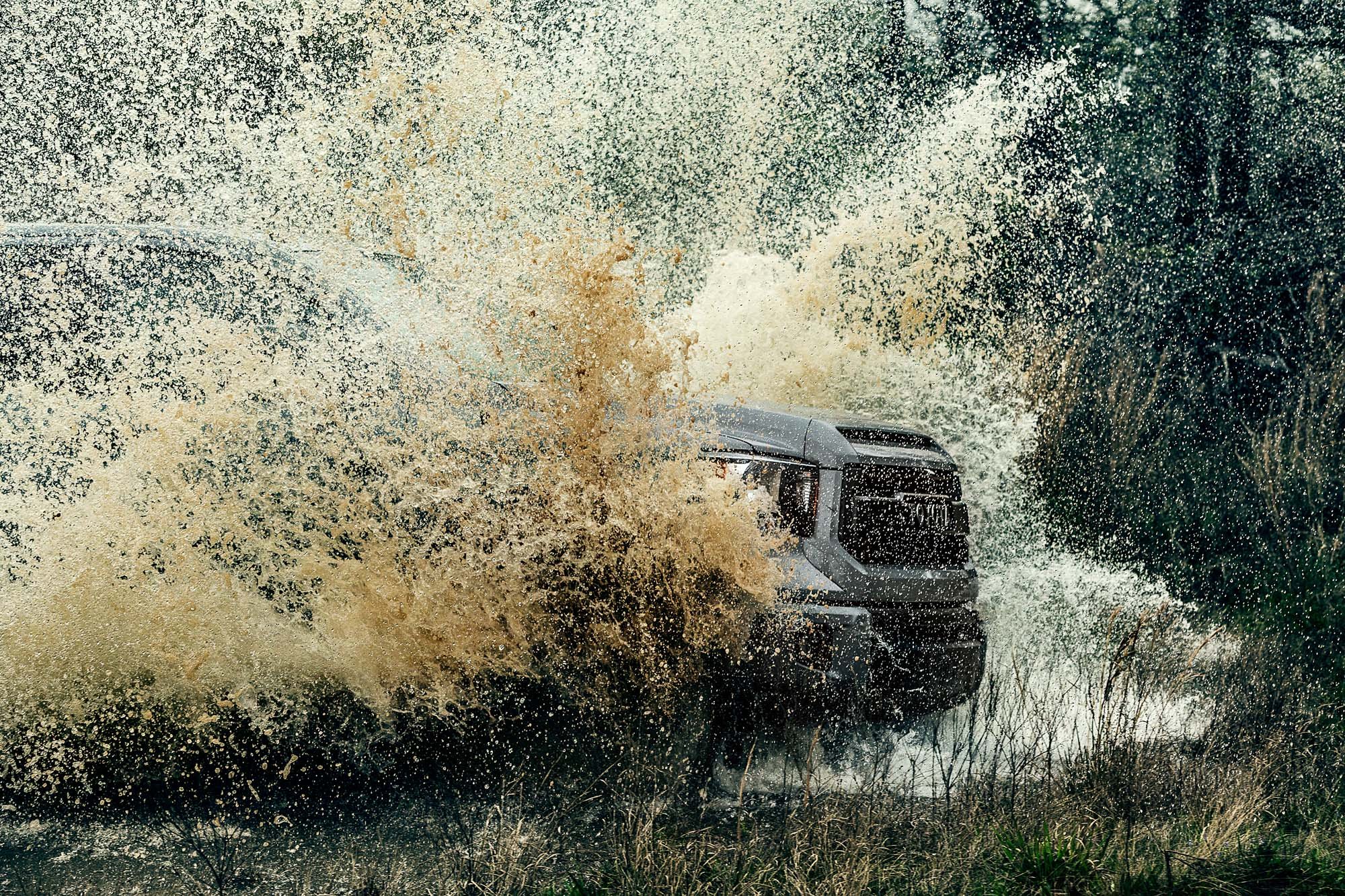 A black truck driving through a flooded area, creating large splashes of water around it amidst trees and tall grass.