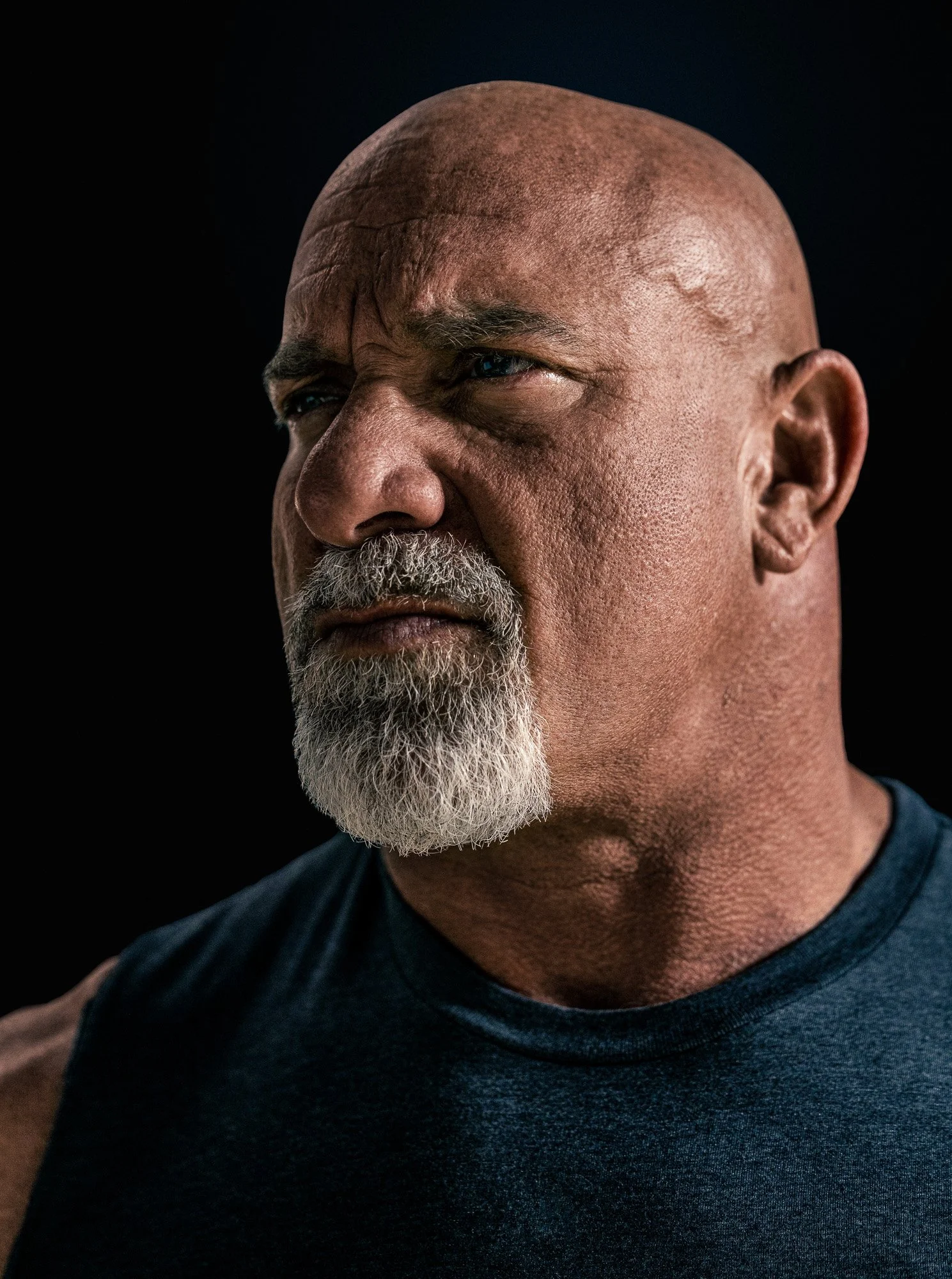Close-up portrait of an older African American man with a white beard, wearing a dark sleeveless shirt, looking thoughtfully into the distance against a black background.