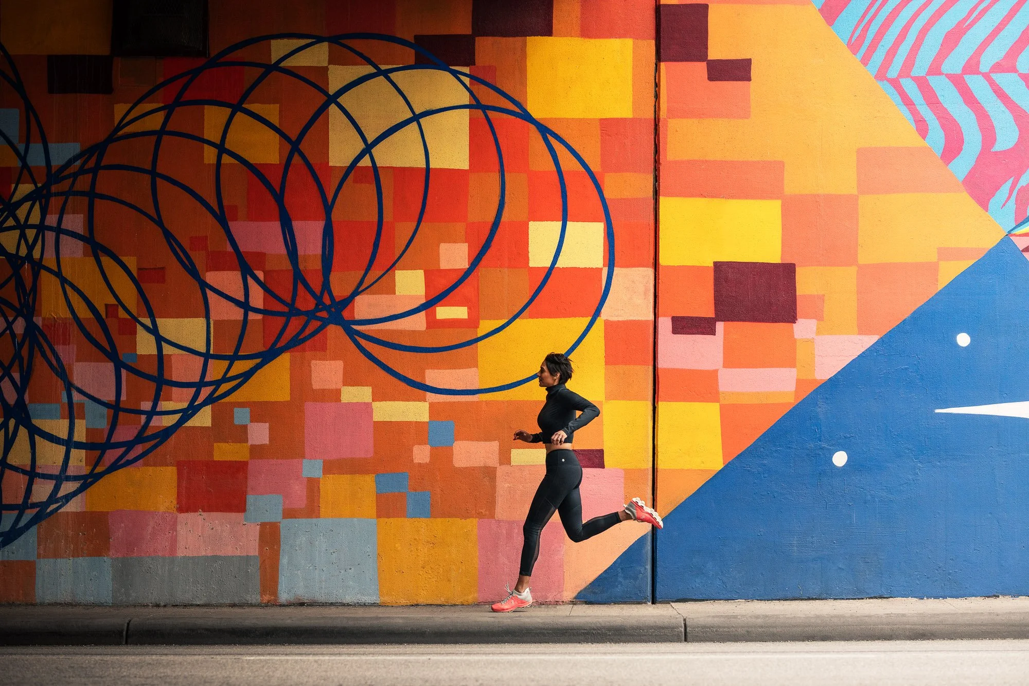 A woman jogging past a colorful mural painted on a wall, featuring geometric shapes and vibrant colors including red, yellow, blue, and pink, with a pattern of overlapping blue circles in the upper left portion.