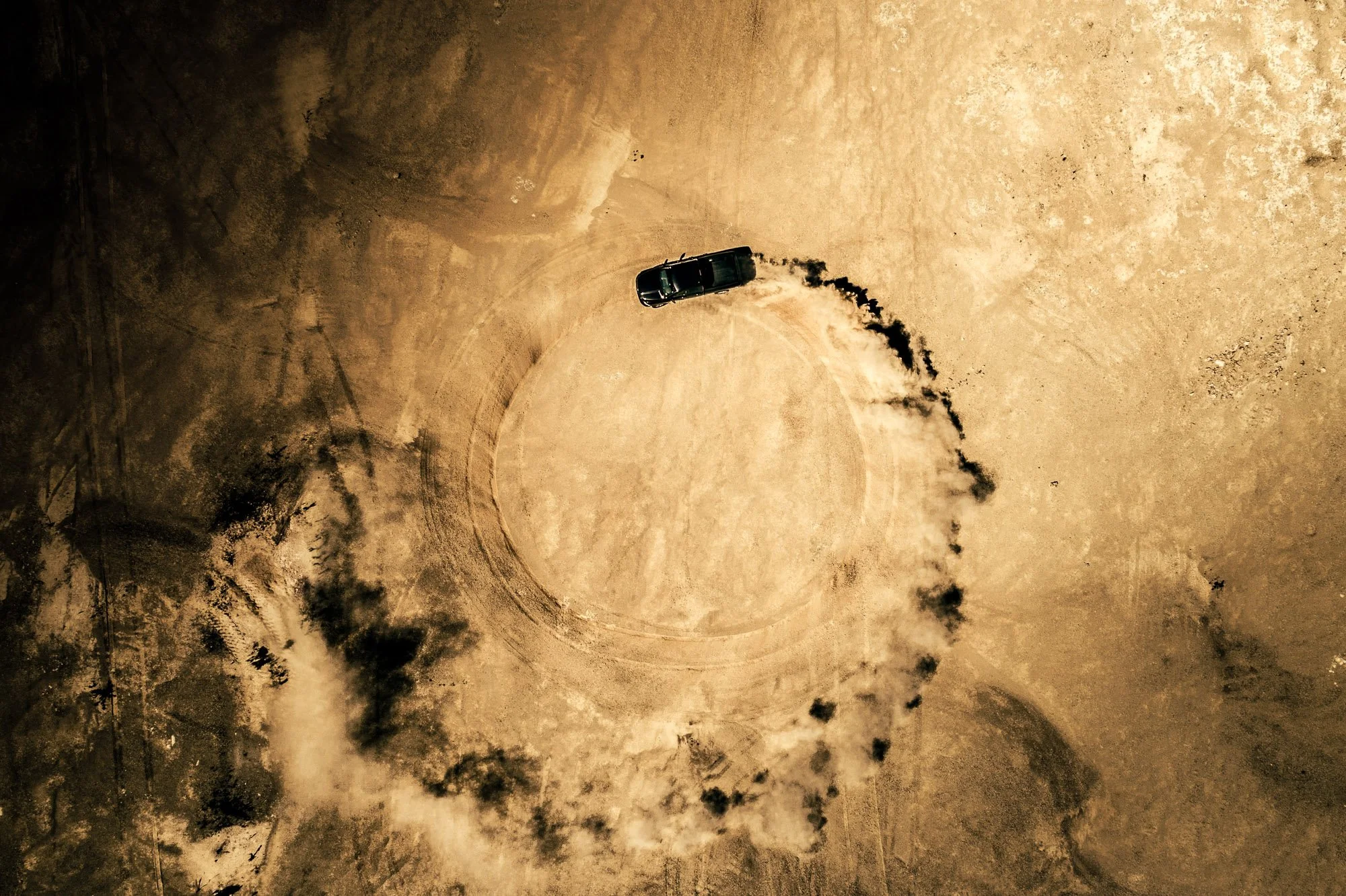 A black car performing a burnout on a dusty, circular dirt area, creating a ring of smoke in an open, arid landscape at night.