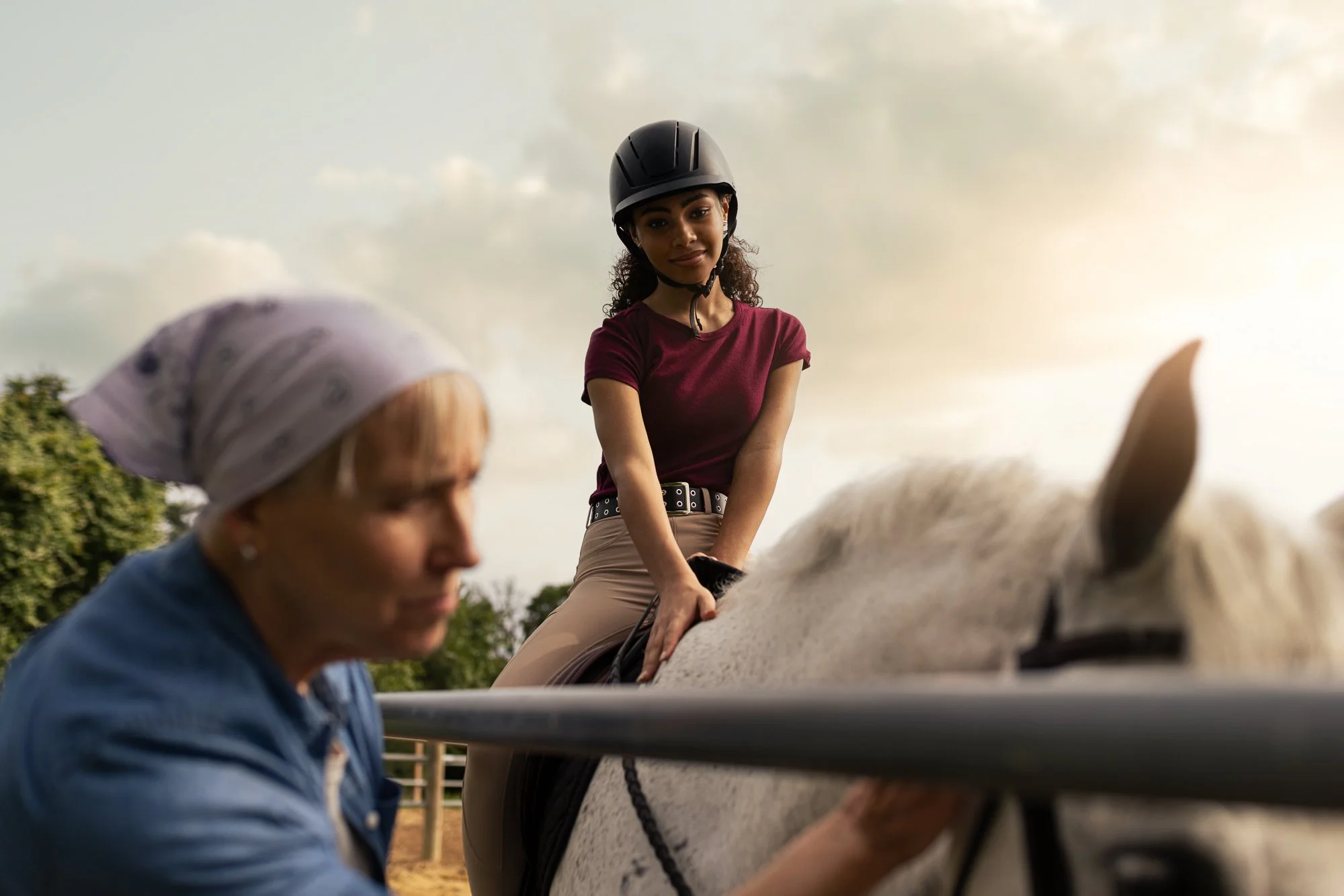 A young woman wearing a helmet sits on a white horse during a riding lesson as an instructor assists her.