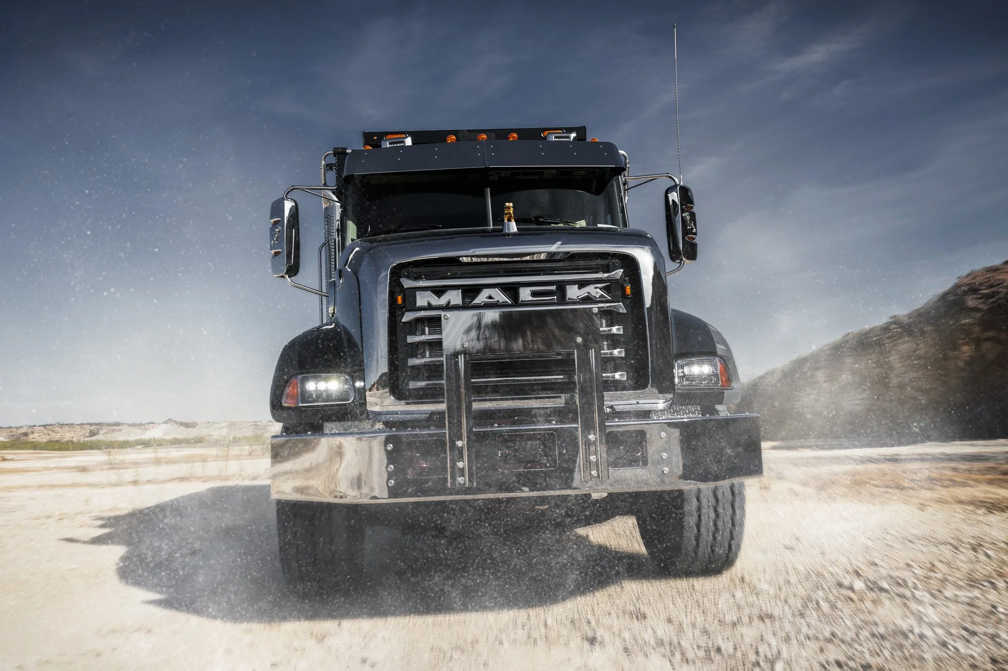 A large black Mack semi-truck driving on a dirt road, kicking up dust with a clear blue sky in the background.