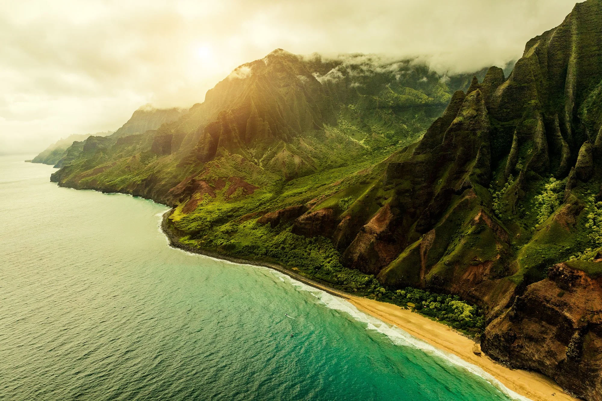 Aerial view of lush green mountains meeting the ocean with a sandy beach at the base, fog rolling over the peaks, and soft sunlight illuminating the landscape.