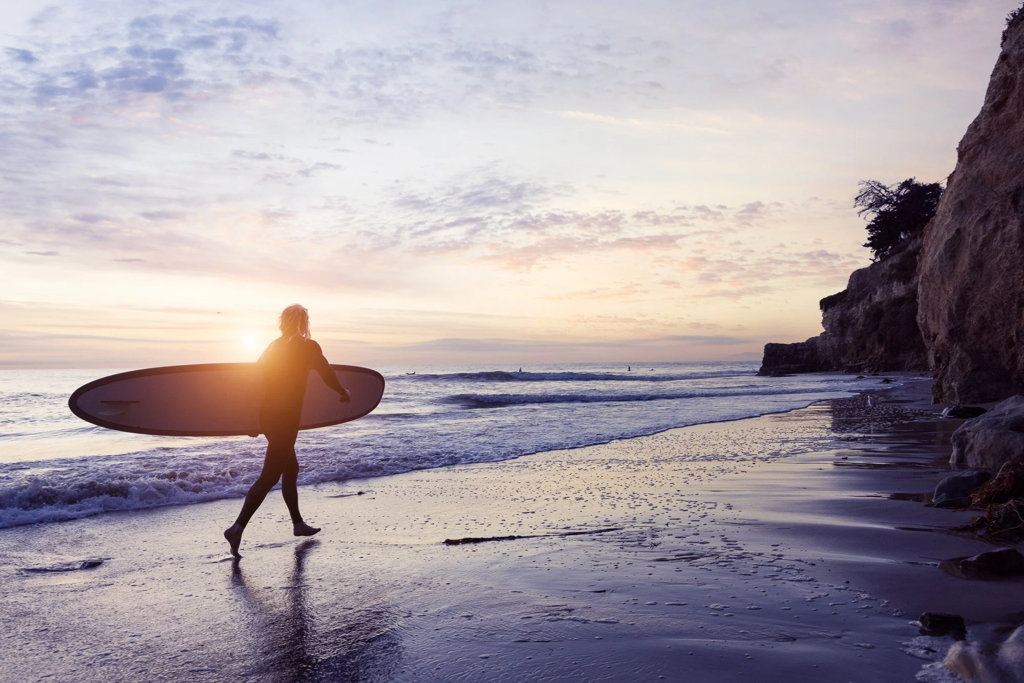 A person holding a surfboard walks along the beach at sunset, with the ocean, waves, and cliffs in the background.