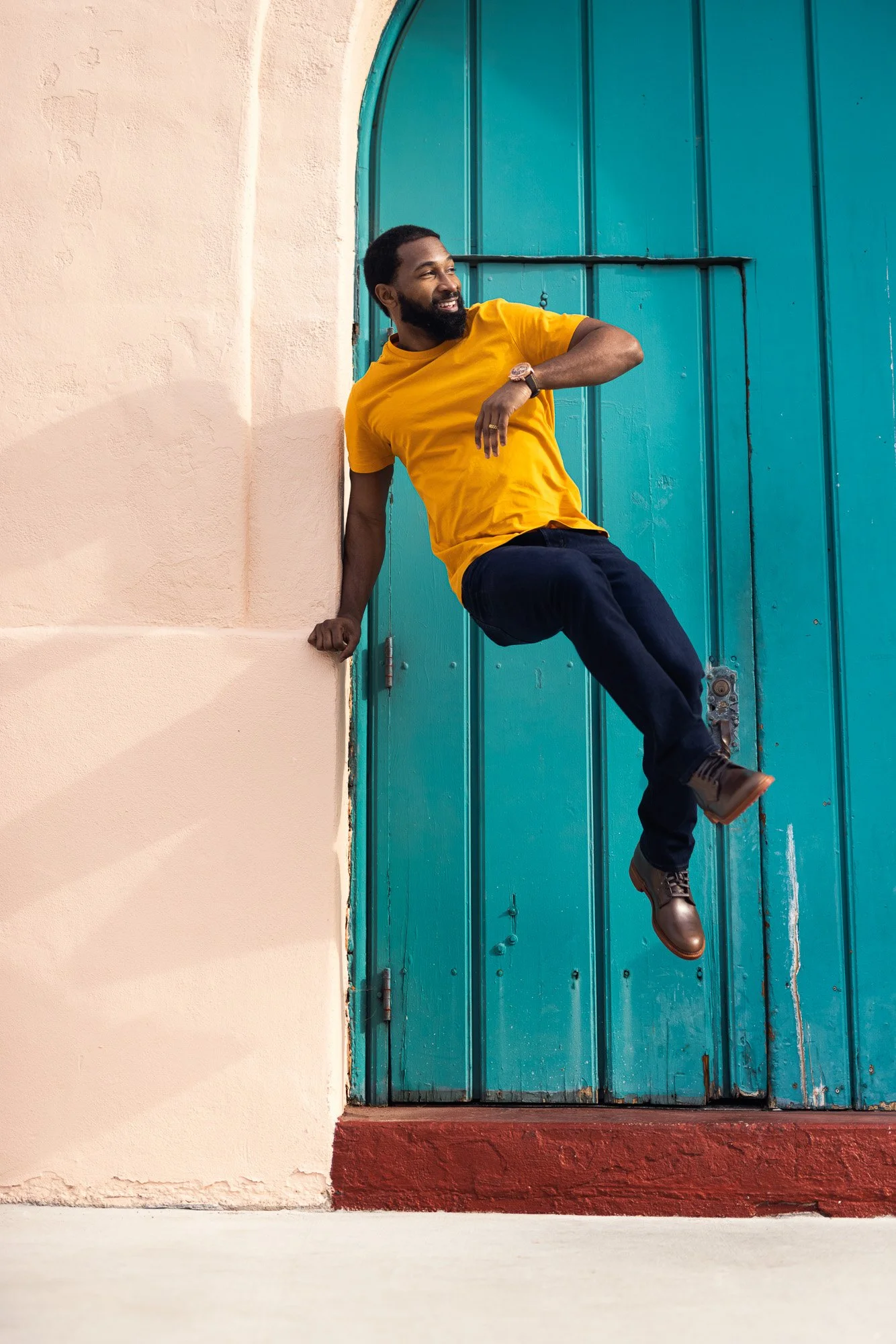 A smiling man with a beard in a yellow t-shirt and dark pants is sitting on a wall, with a turquoise door in the background.