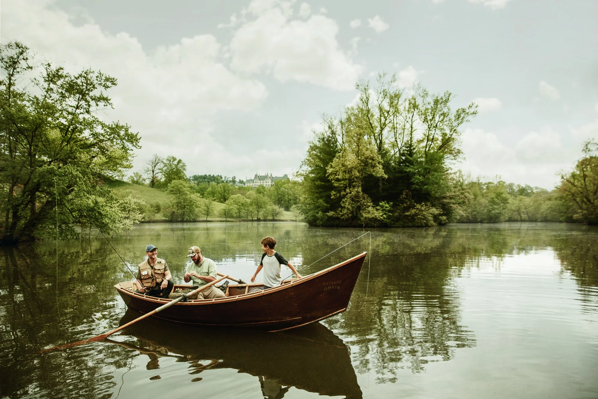 Three people fishing in a small wooden boat on a calm lake surrounded by trees and a cloudy sky.