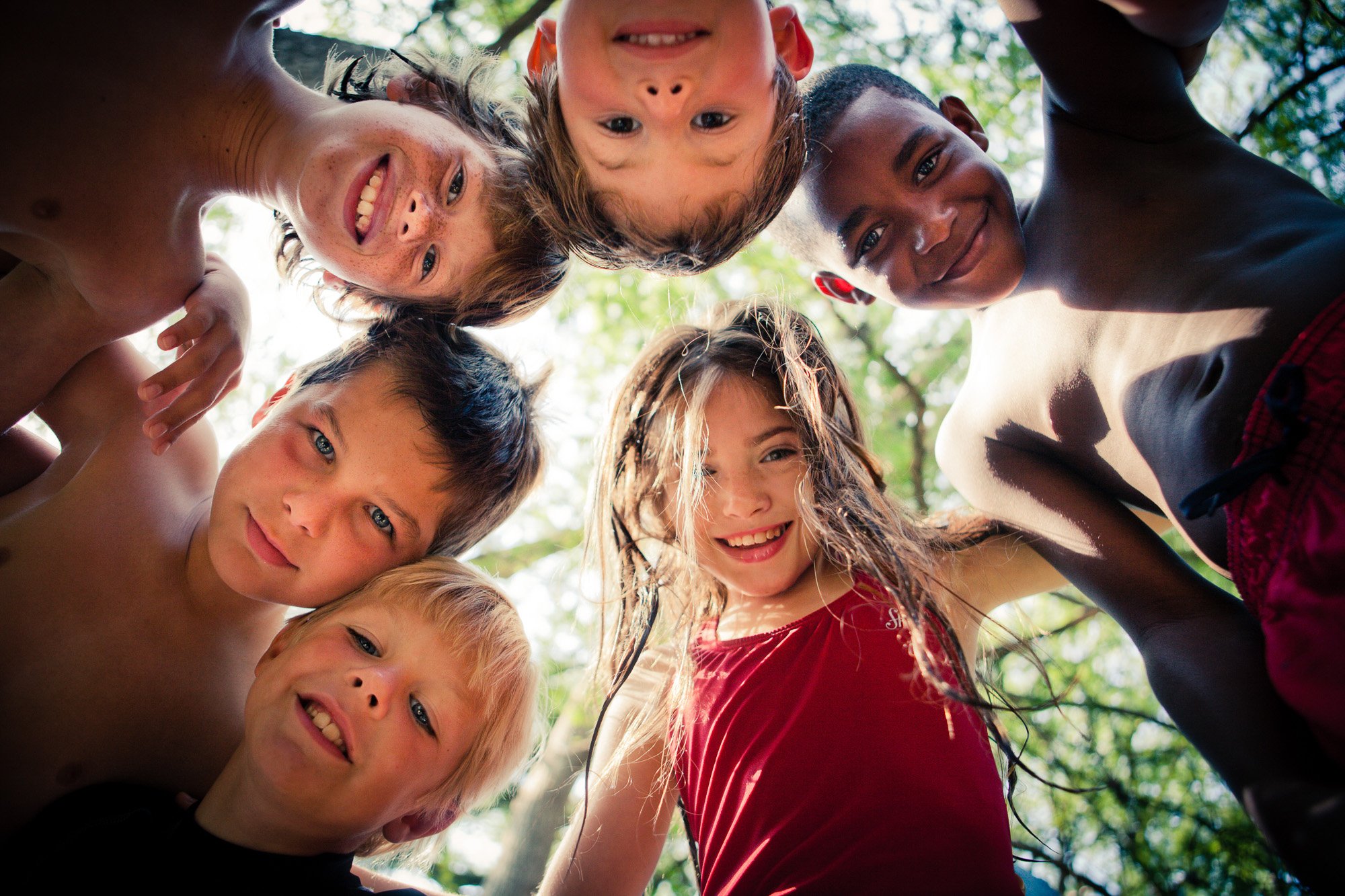 A group of seven children looking down at the camera with smiles, standing outdoors with trees and sunlight in the background.