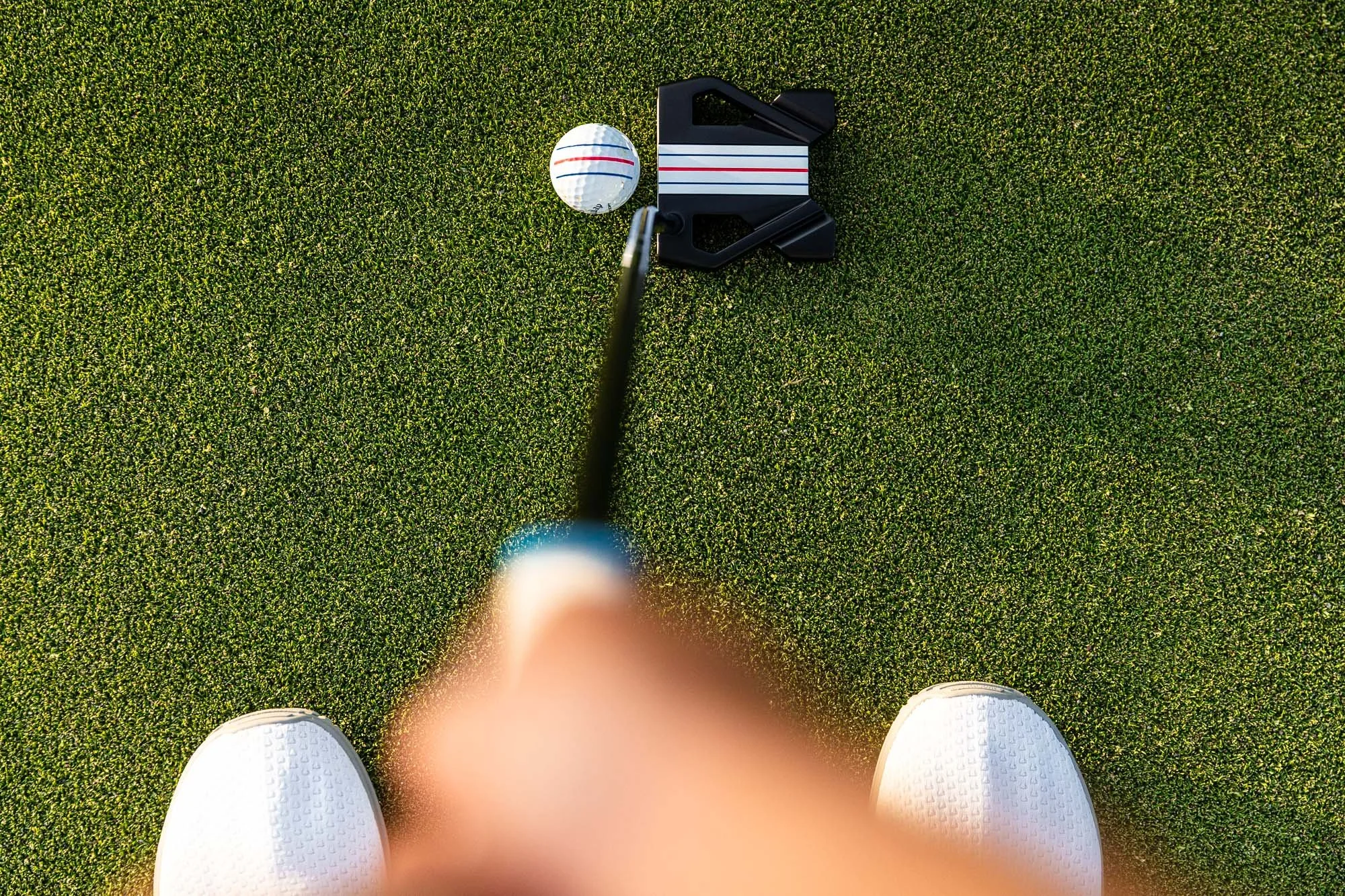 Close-up of a person's feet in white shoes on a golf putting green, viewing a golf ball near a putter and golf marker.