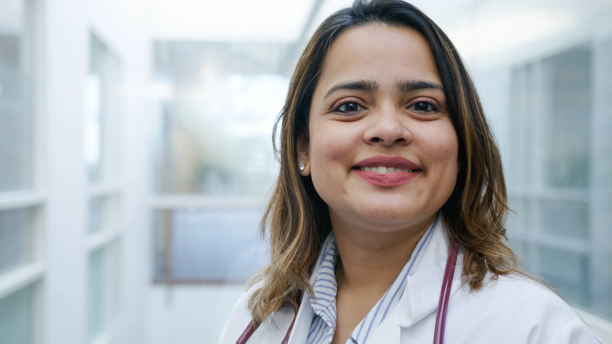 A woman with dark brown hair and light skin, smiling, wearing a white coat and a stethoscope around her neck, in a bright indoor environment.