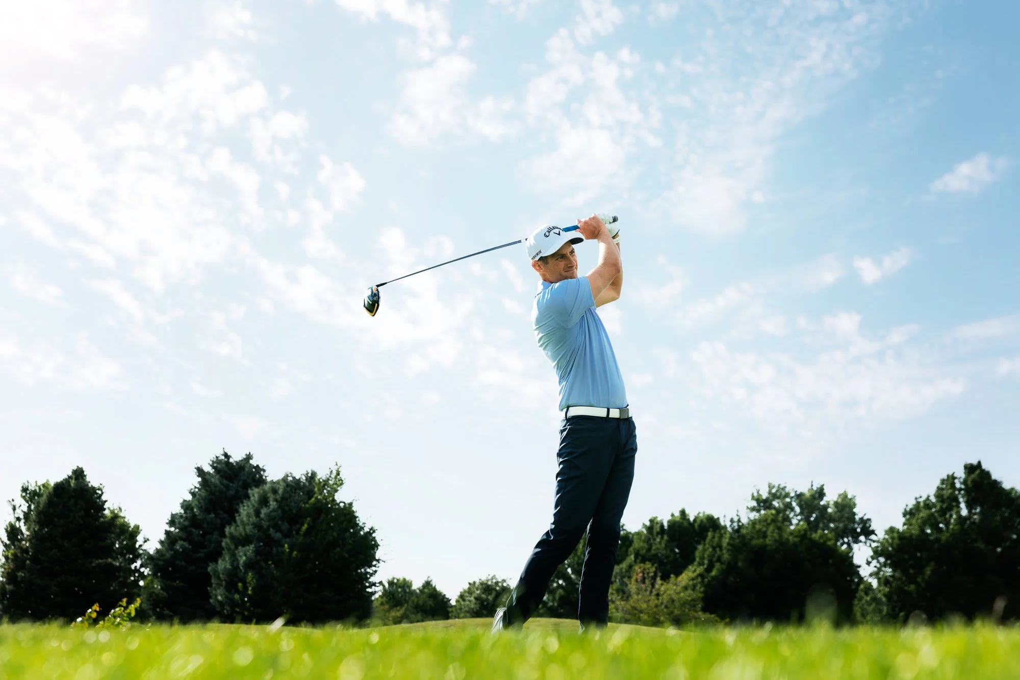 A man swinging a golf club on a golf course with blue sky and trees in the background.