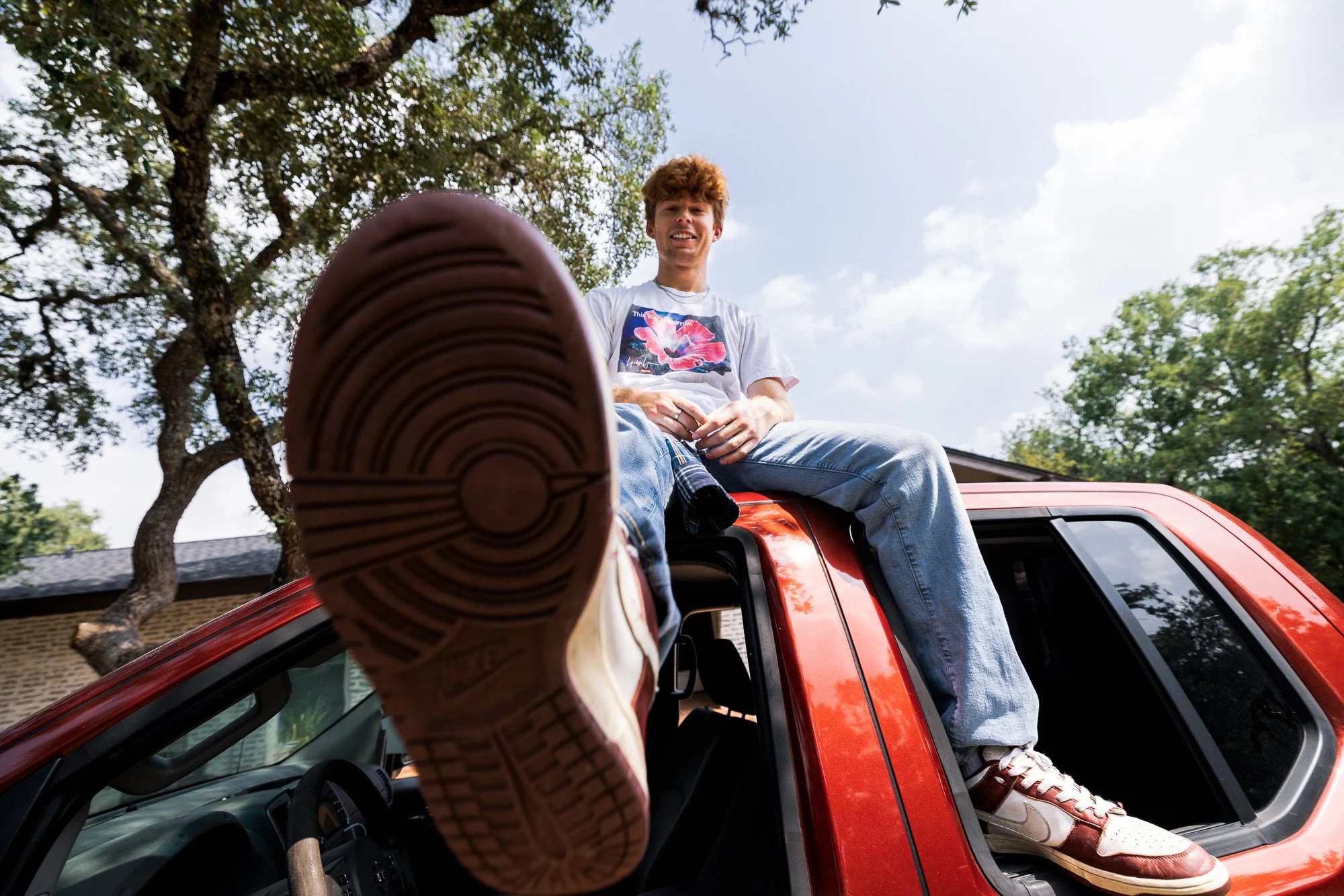 Young man sitting on the roof of a red truck with a foot extended toward the camera, outdoors with trees and a partly cloudy sky