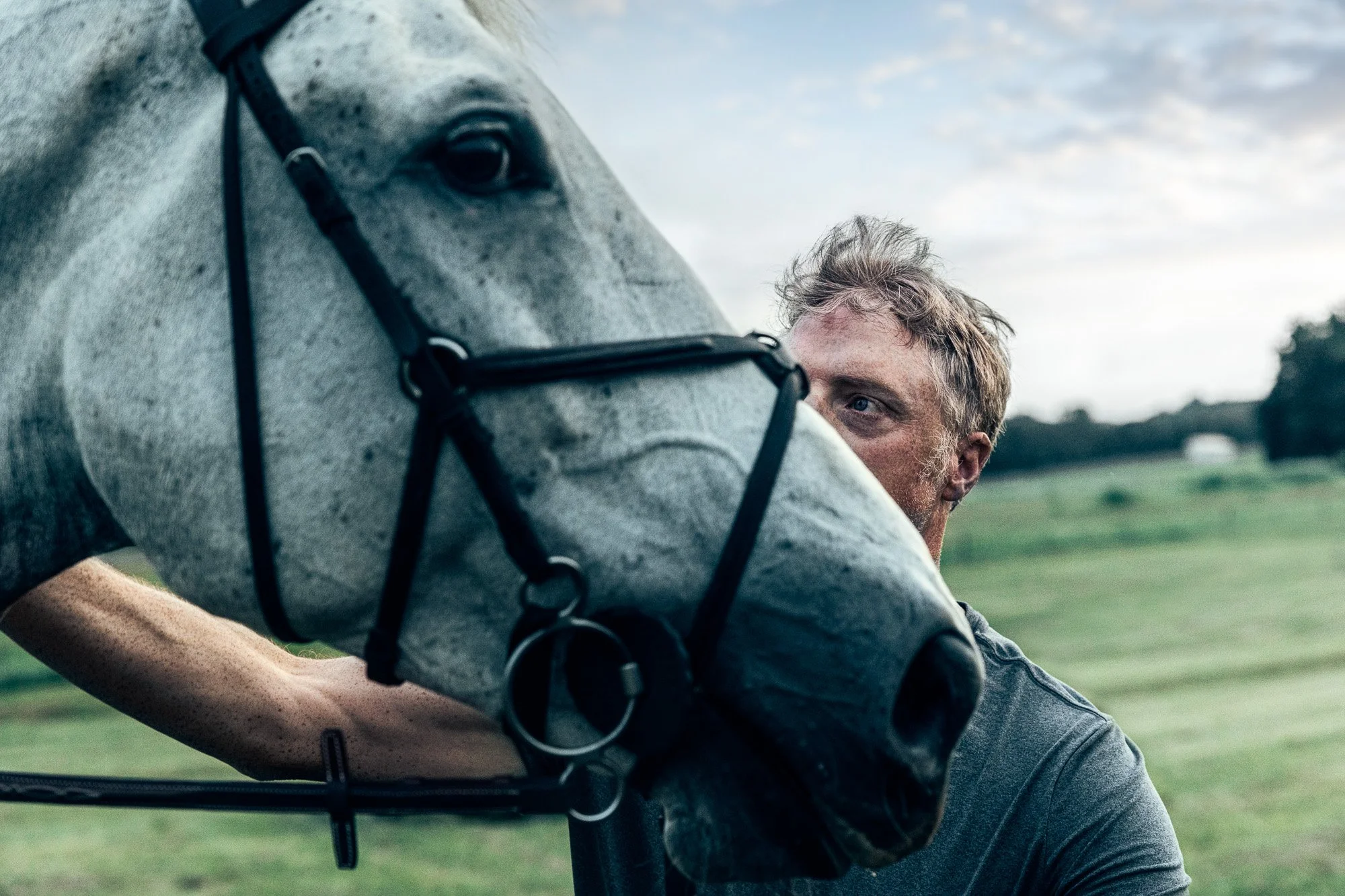 A man with short, messy hair looking at a gray horse with a black bridle outdoors on a cloudy day.