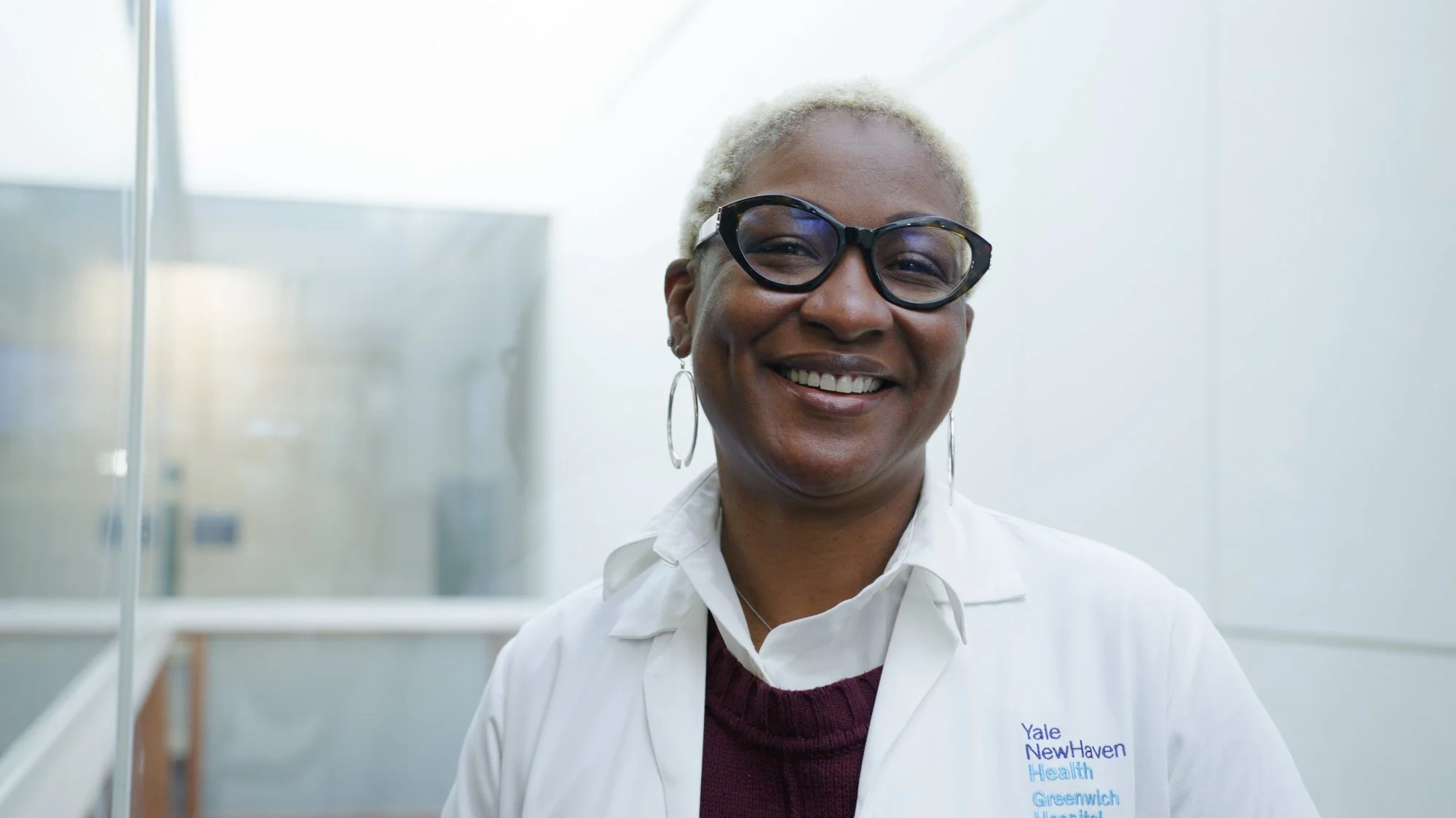 A smiling African American woman with short blonde hair and glasses, wearing a white lab coat at Yale New Haven Health, standing in a medical or hospital setting.