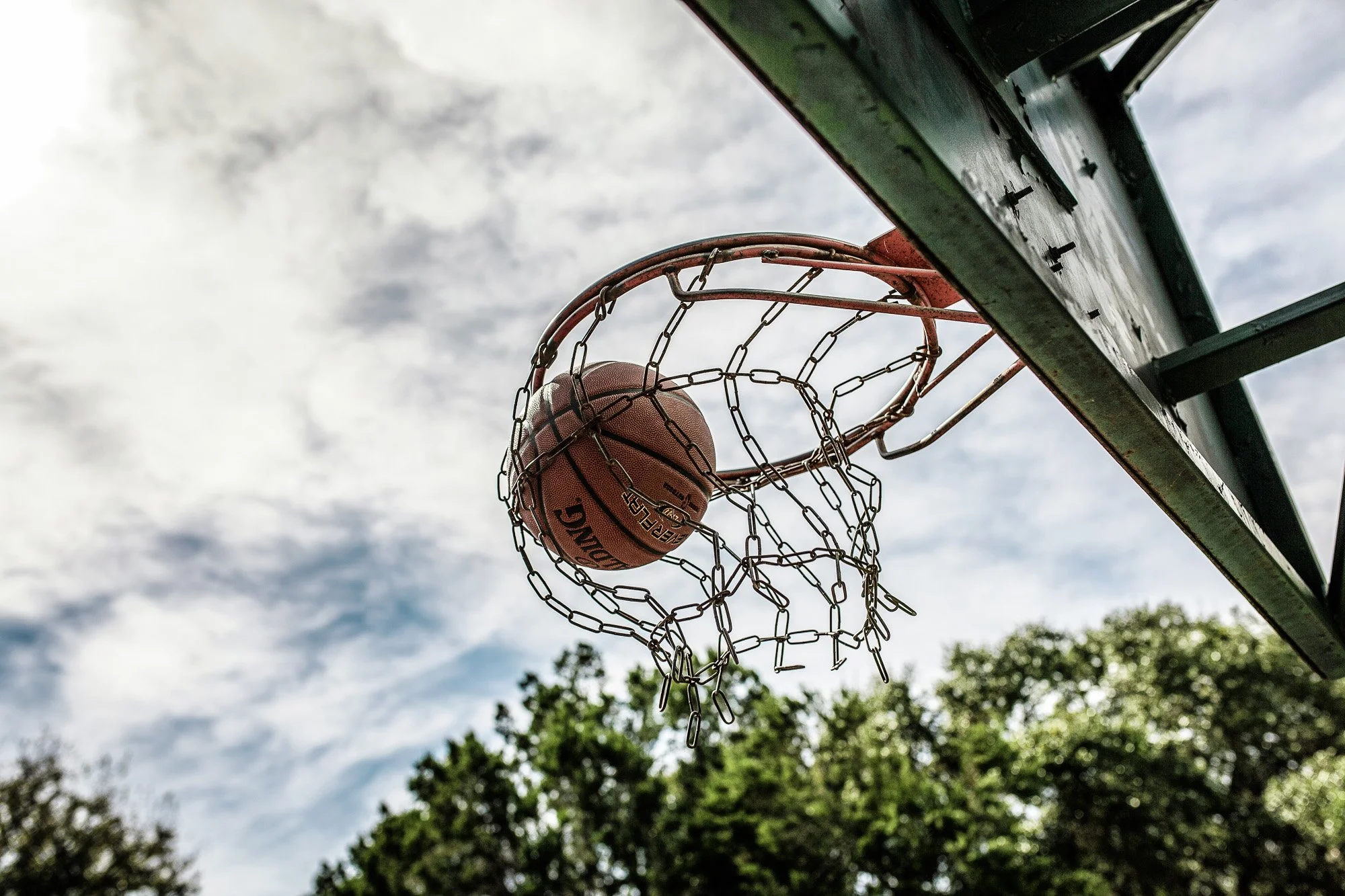 A basketball shot is made through the net on an outdoor basketball hoop against a partly cloudy sky with trees in the background.