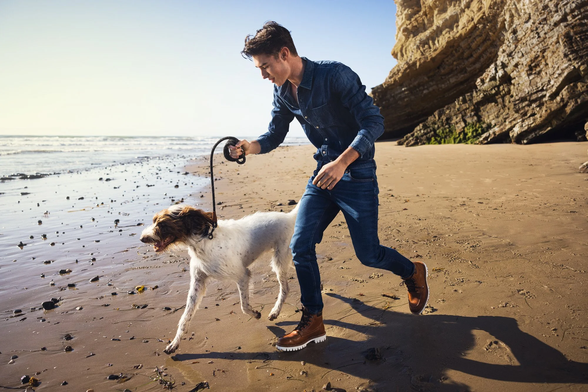 A young man in jeans and a denim shirt playing with a dog on a sandy beach with a rocky cliff in the background.